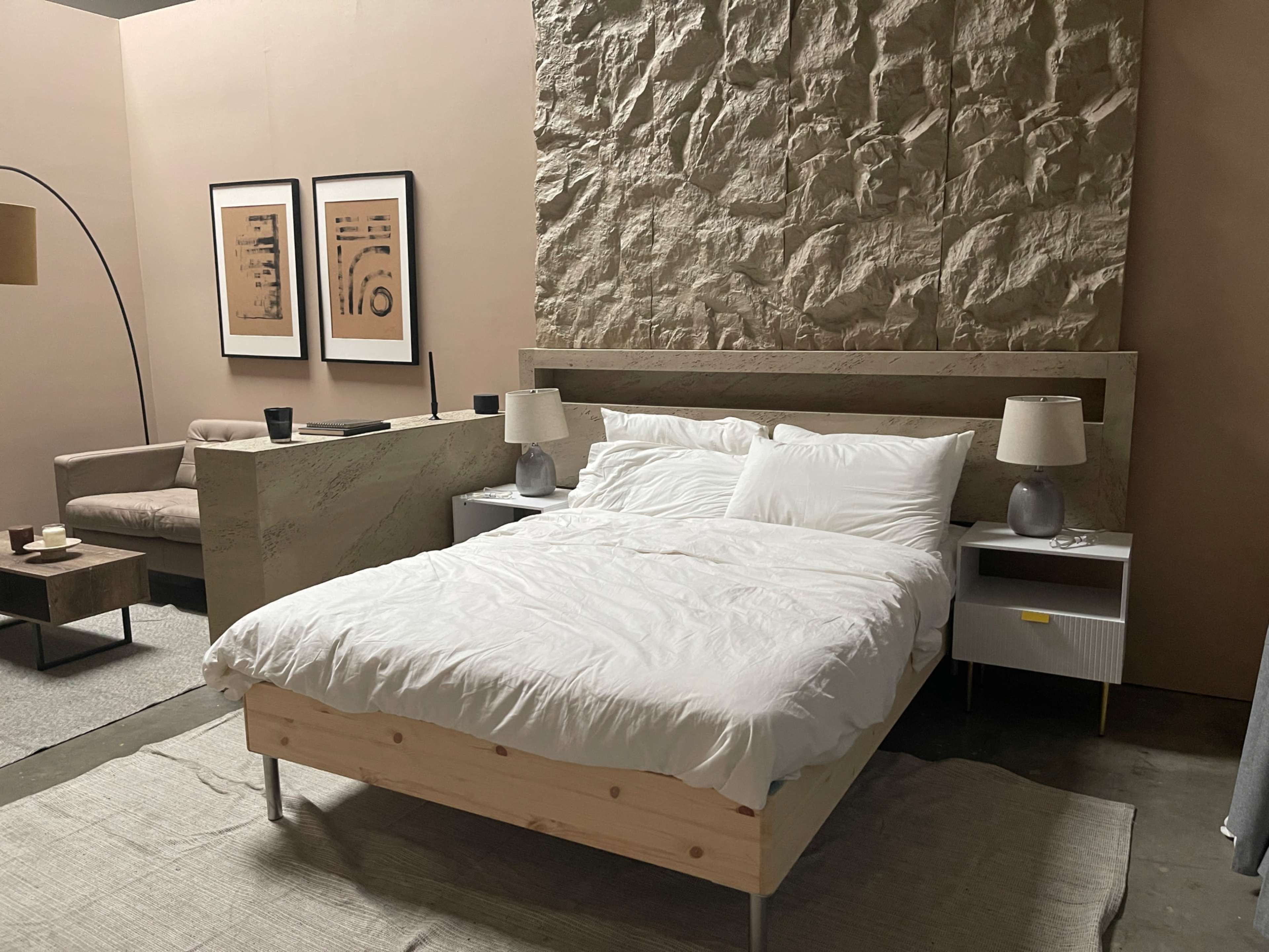 A modern bedroom with a wooden bed, two nightstands, a textured stone wall, and a minimalist couch arranged in the space.