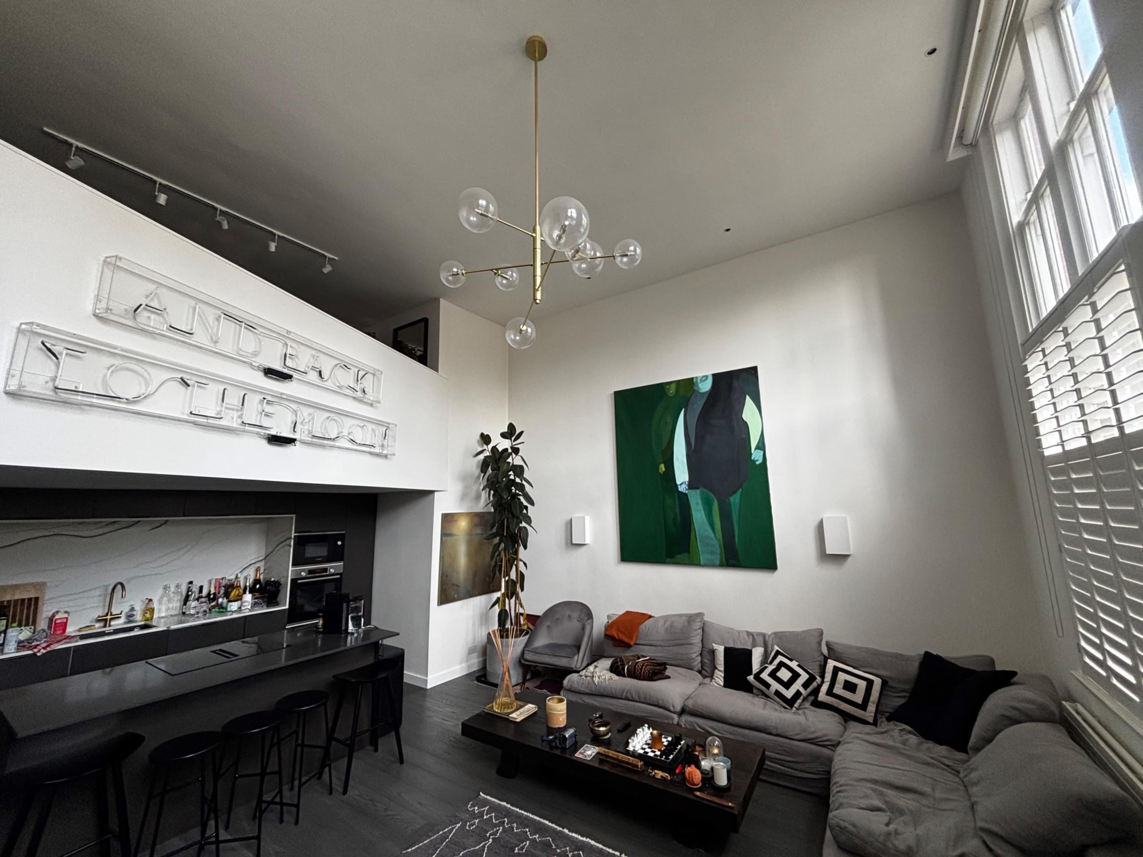 The image shows a modern living room with high ceilings, a gray sofa, an abstract painting on the wall, and a minimalist kitchen area with bar stools.