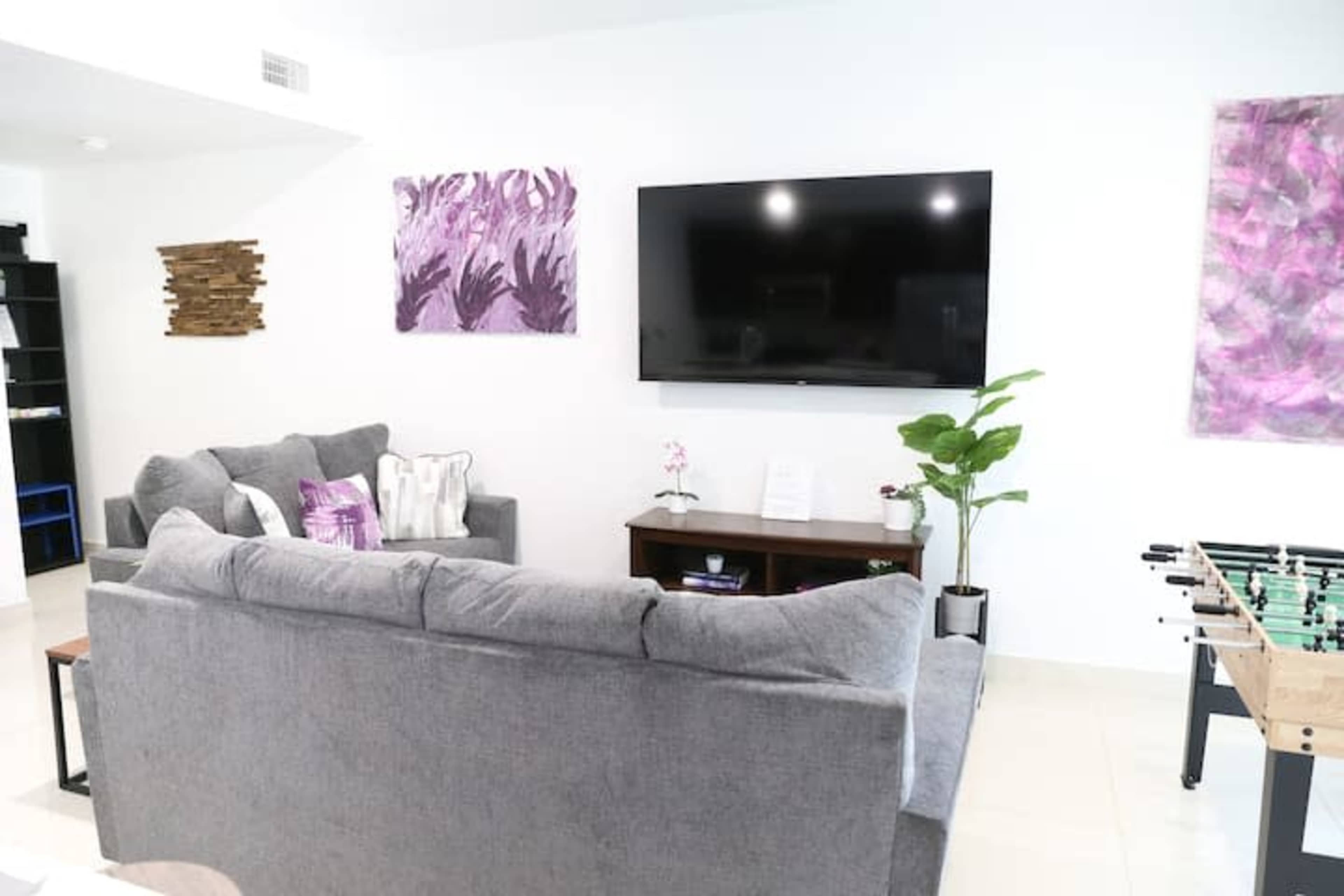 A modern living area features a gray sectional sofa, a wall-mounted TV, and decorative purple artwork, with a foosball table in the corner.