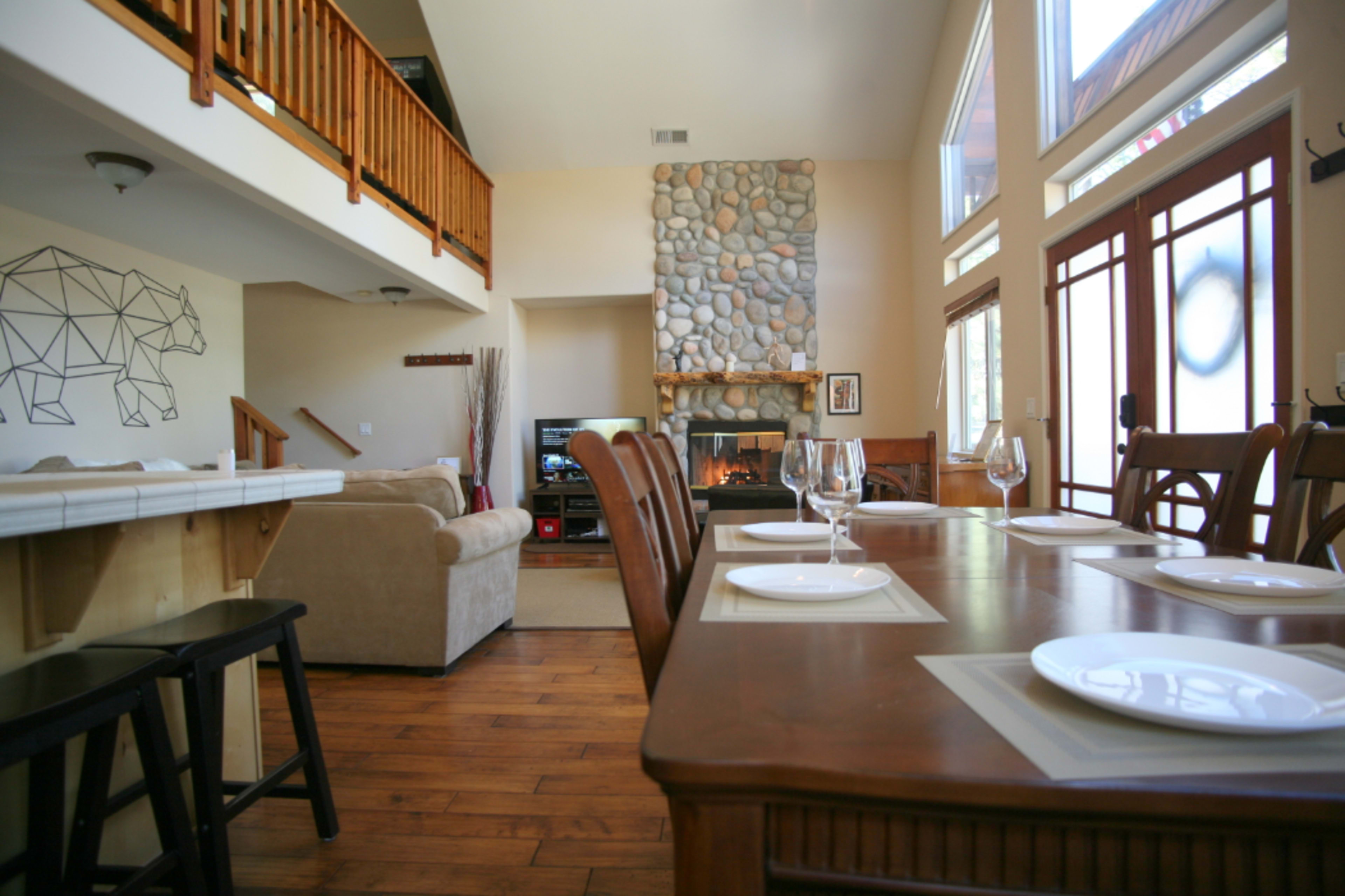 The image shows a spacious living area with a dining table set for a meal, a large stone fireplace, and a view of a comfortable seating area with a sofa.