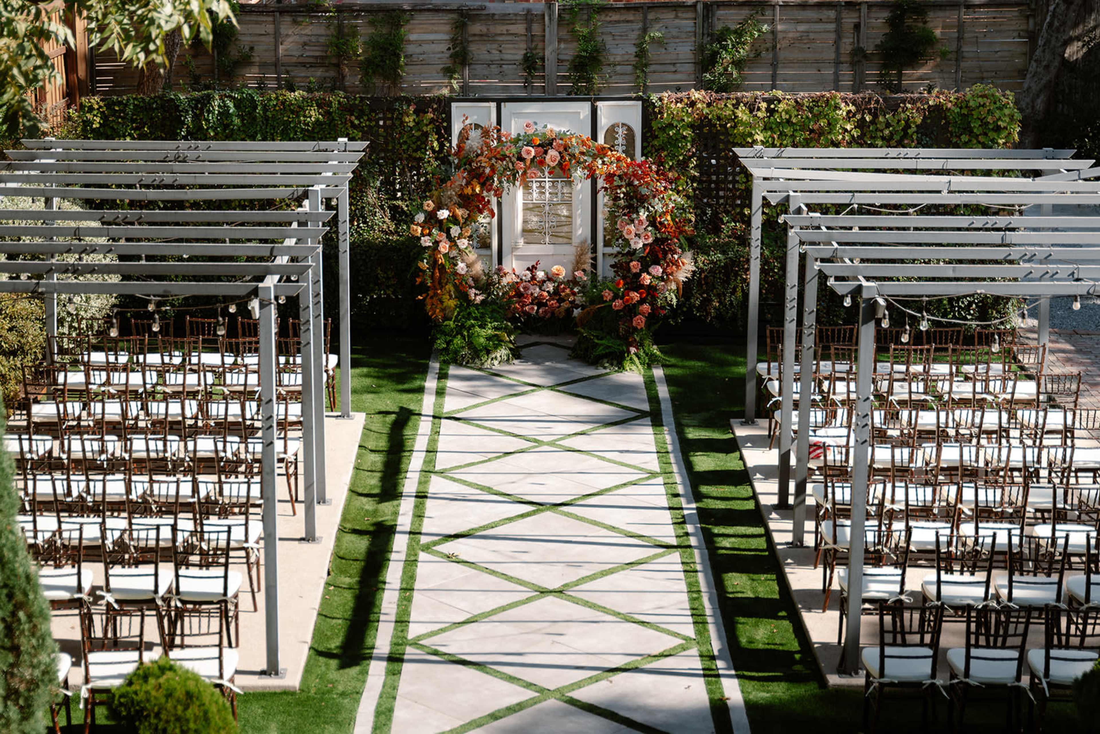 The image shows a wedding ceremony setup with rows of chairs arranged along a paved walkway leading to a floral arch at the front.