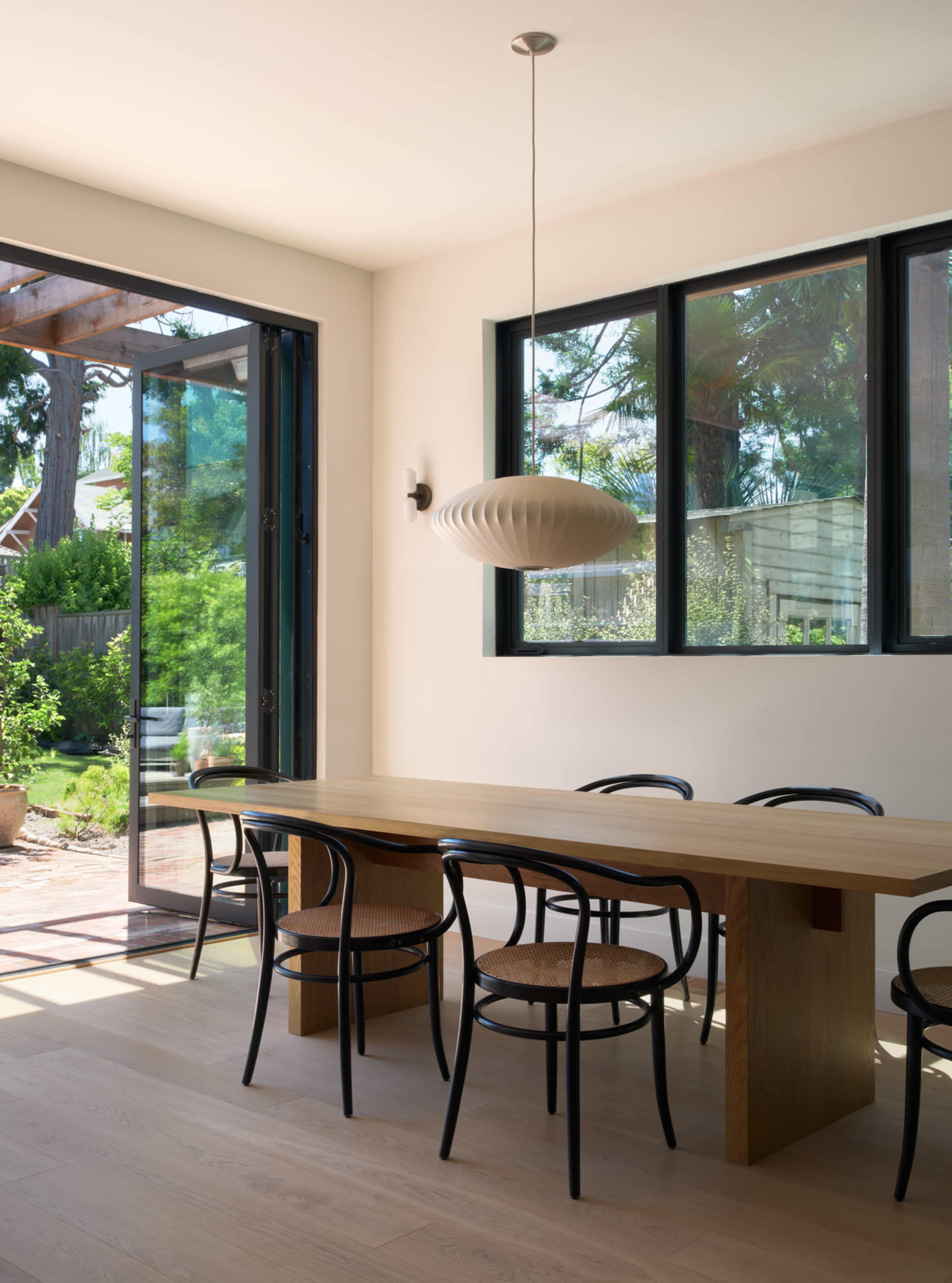 A modern dining area features a long wooden table surrounded by black chairs, with large windows and doors leading to an outdoor garden.