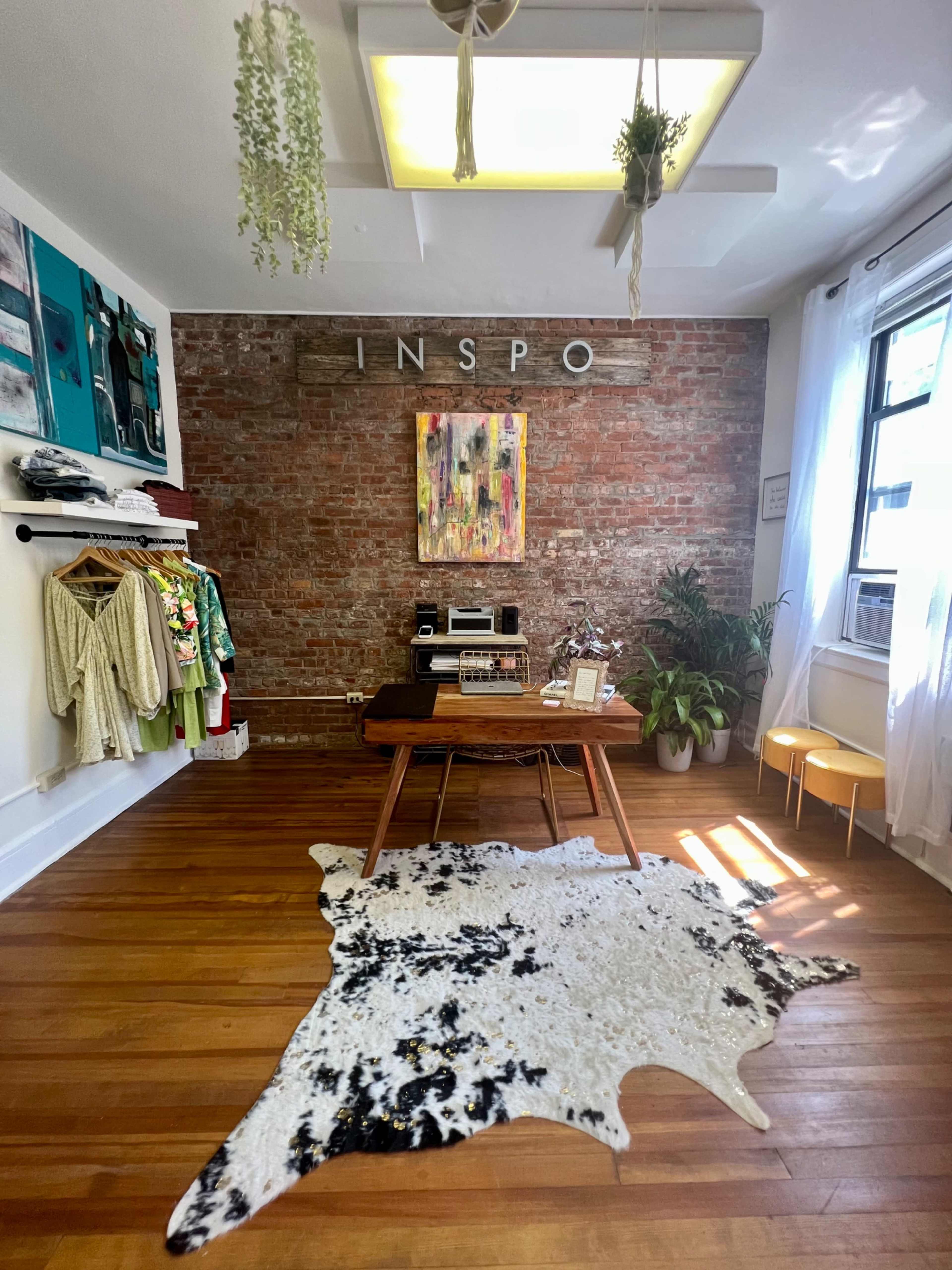 A cozy workspace featuring a wooden table, colorful wall art, a brick wall, clothing displays, and a decorative cowhide rug on polished wooden floors.