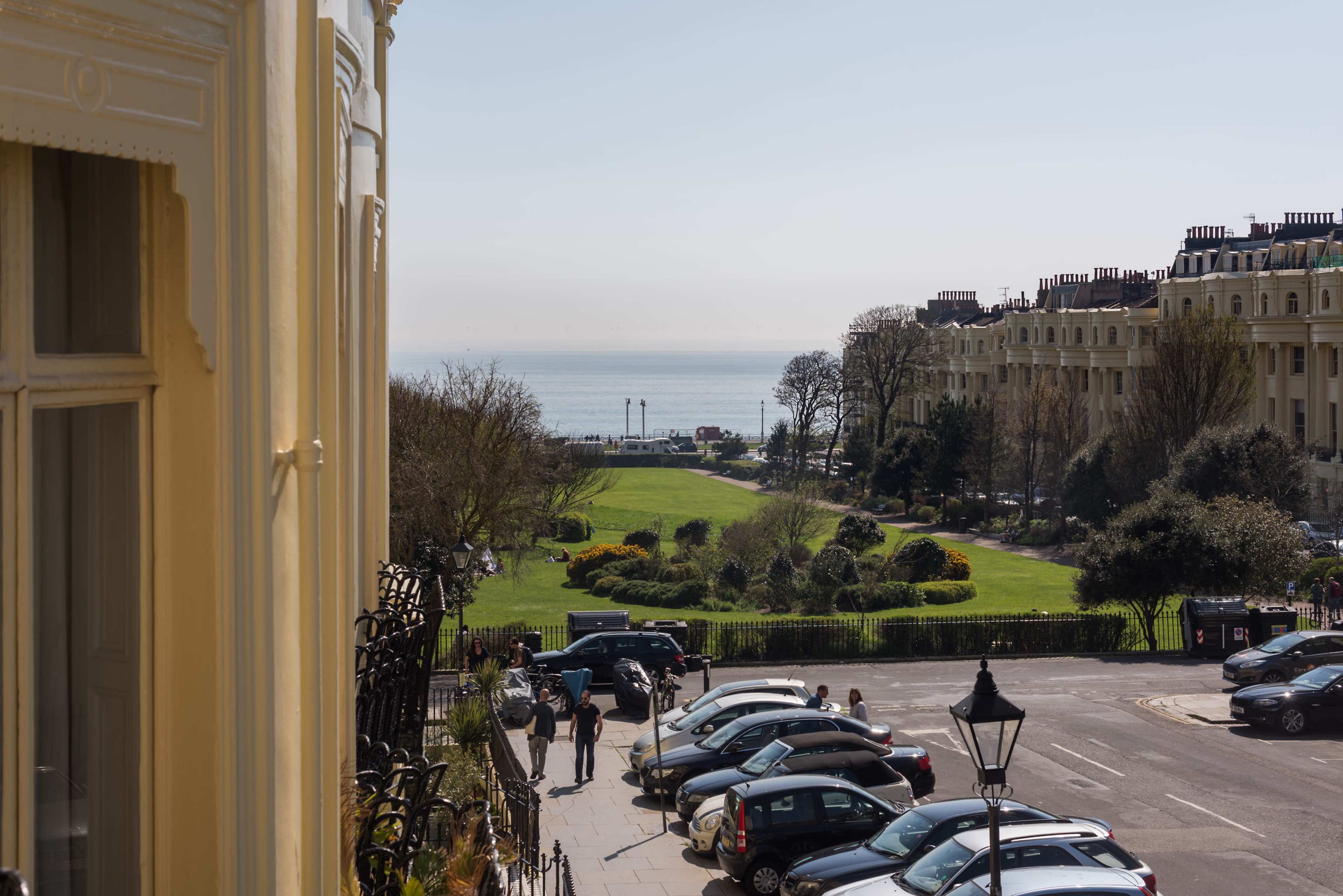 A seaside view with a park, cars parked in the foreground, and buildings lining the street under a clear sky.
