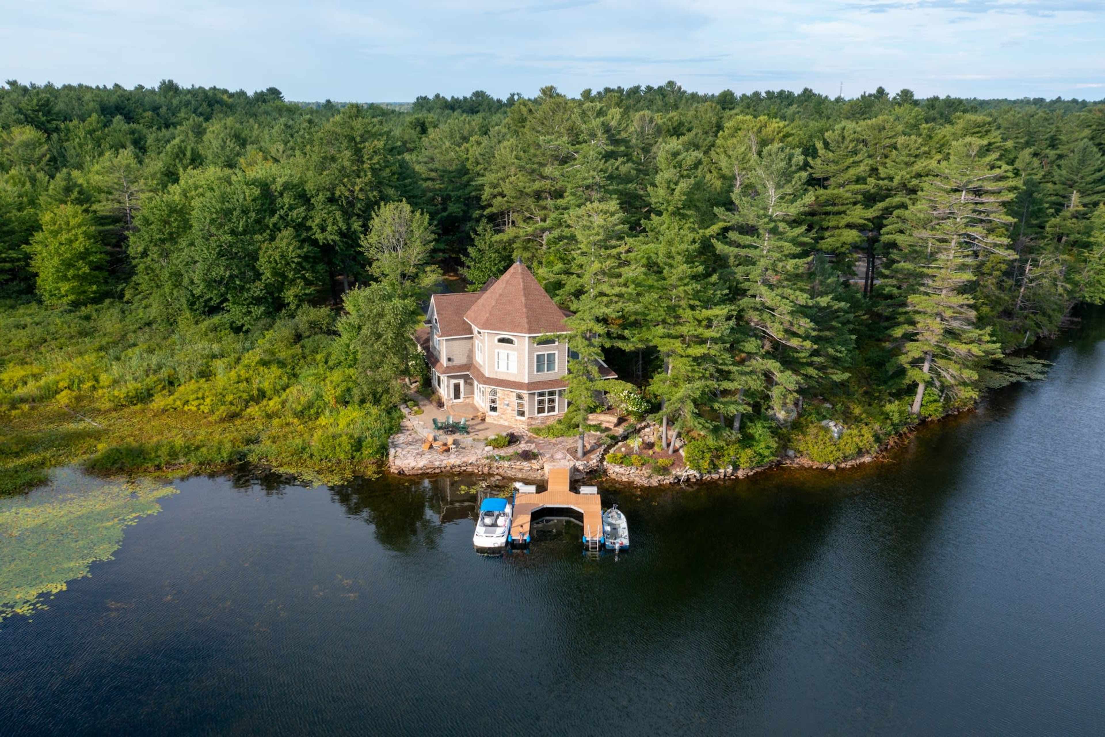 The image shows a lakeside house surrounded by trees, with a dock and two boats by the water.
