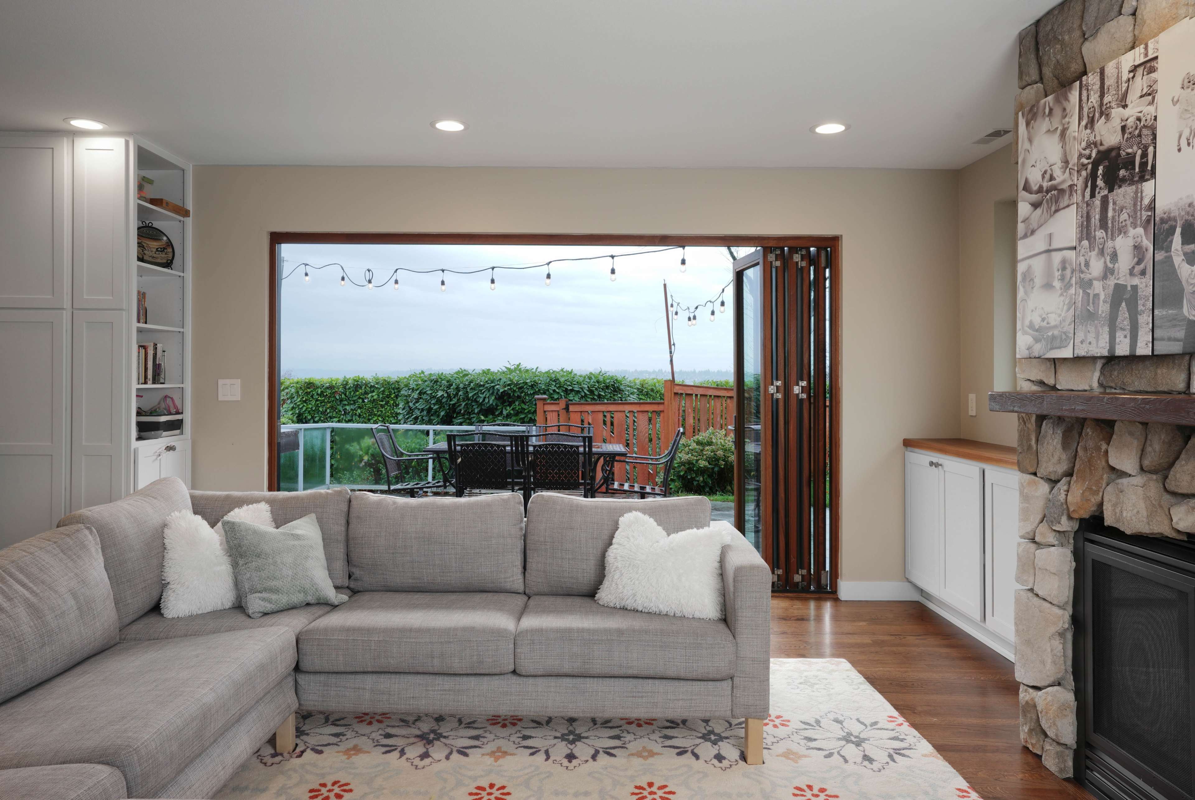 A modern living room features a gray sectional sofa, a stone fireplace, and an open sliding door revealing a view of an outdoor deck and greenery.