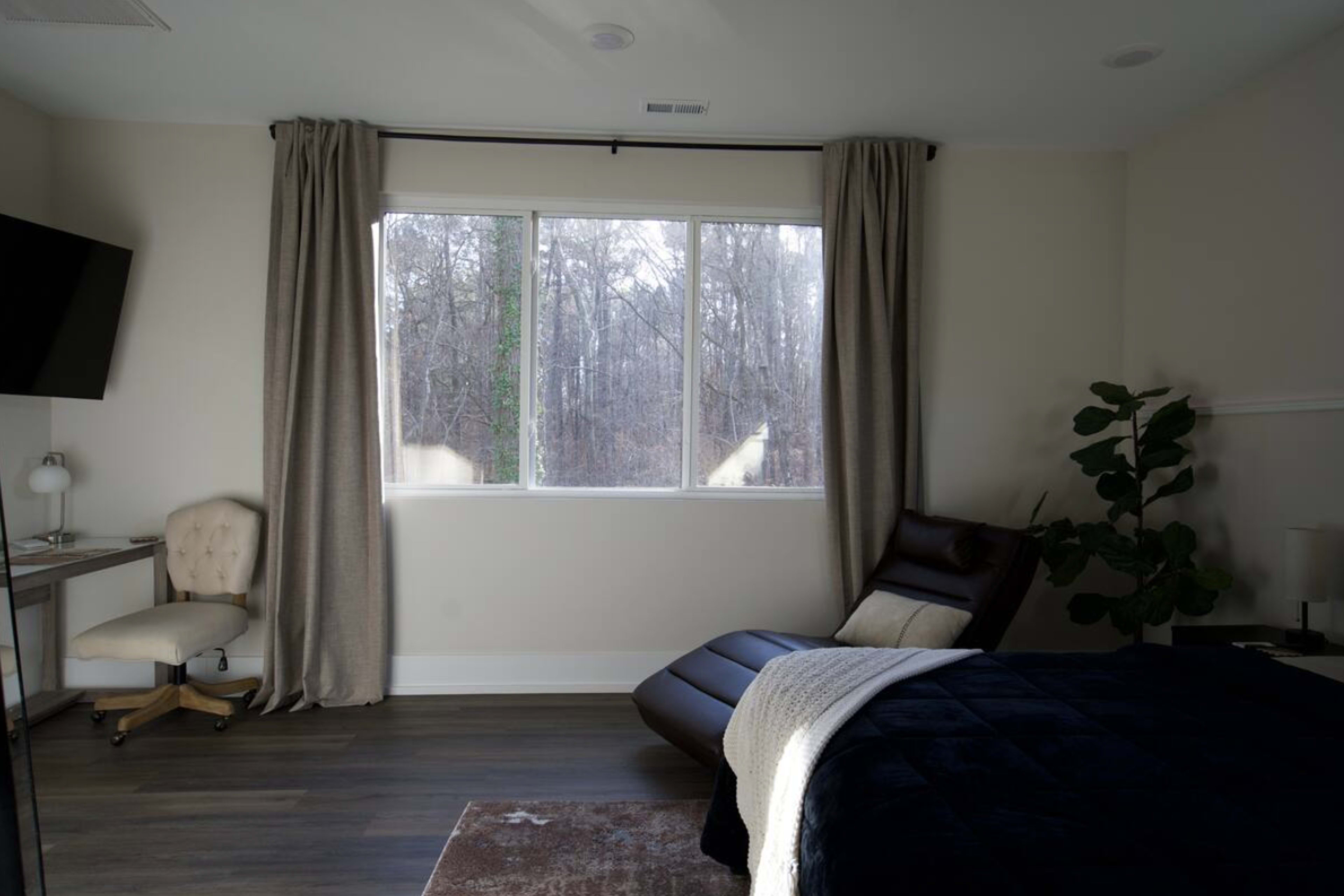A modern bedroom features a brown reclining chair, a bed with a dark duvet, a desk with a lamp, and large windows overlooking a forested area.
