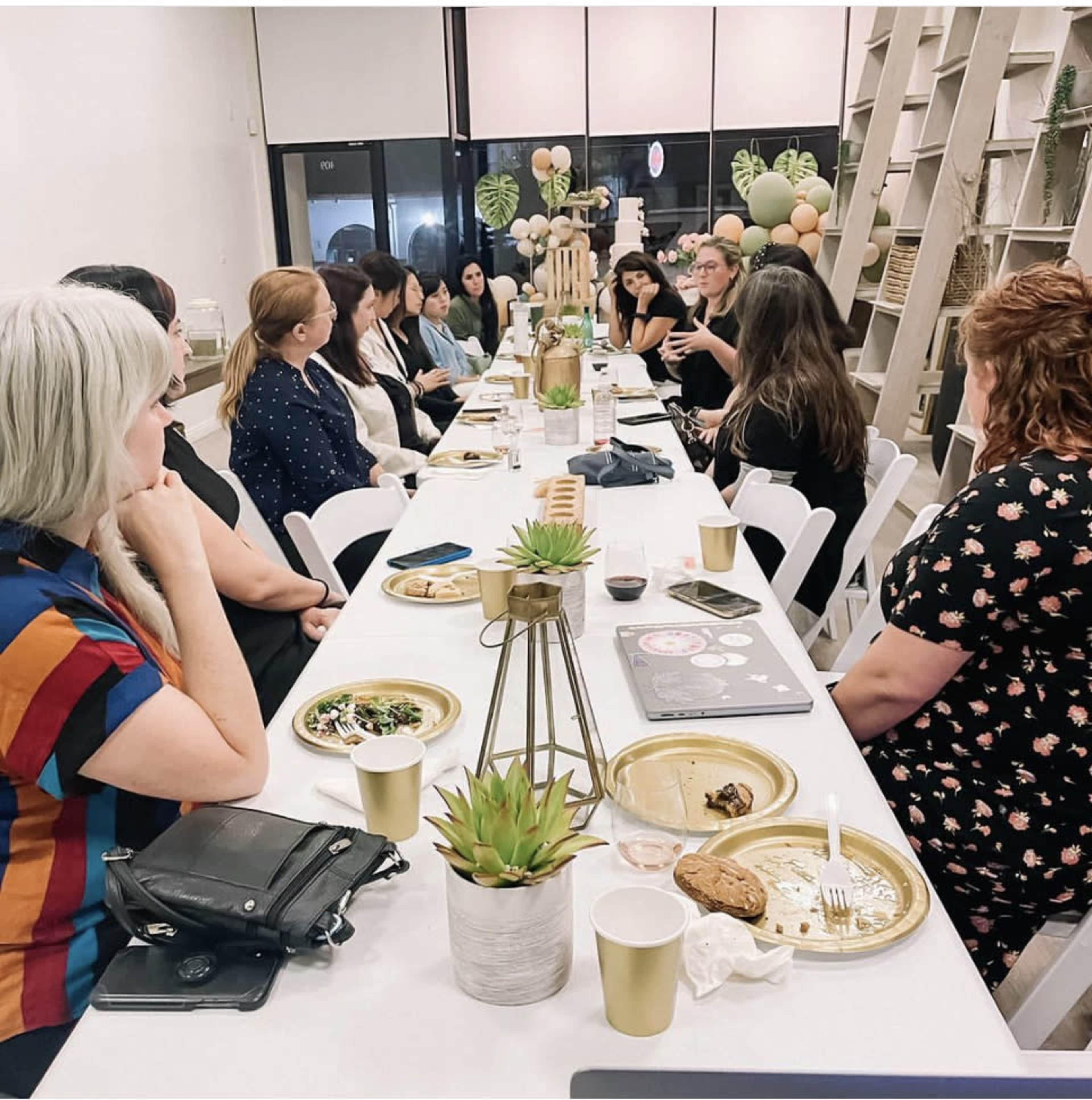 A group of women sits at a long table sharing a meal in a well-lit space decorated with plants and balloons.