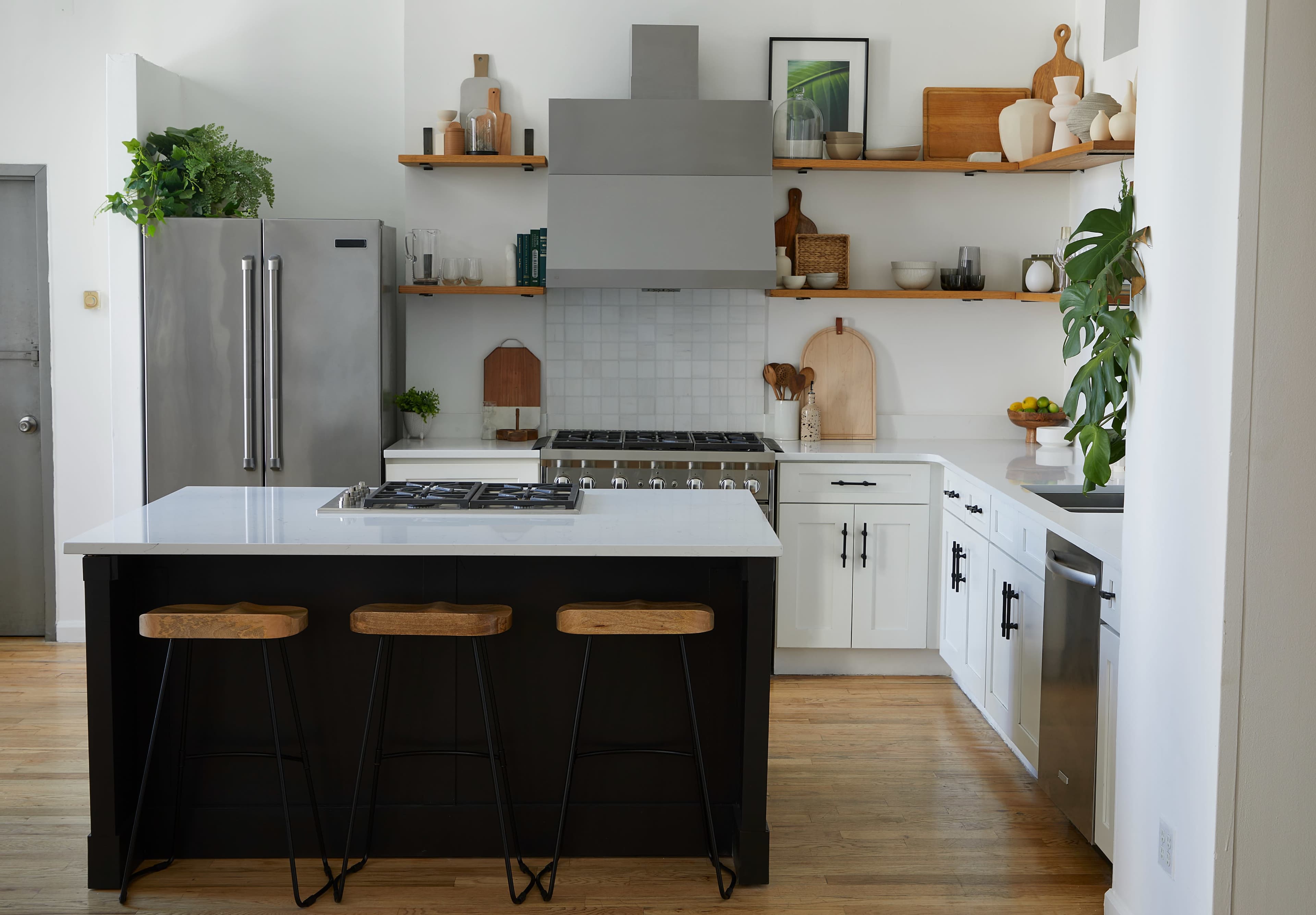 A modern kitchen features a central island with three wooden stools, stainless steel appliances, open shelving displaying various kitchen items, and a combination of white and black cabinetry.