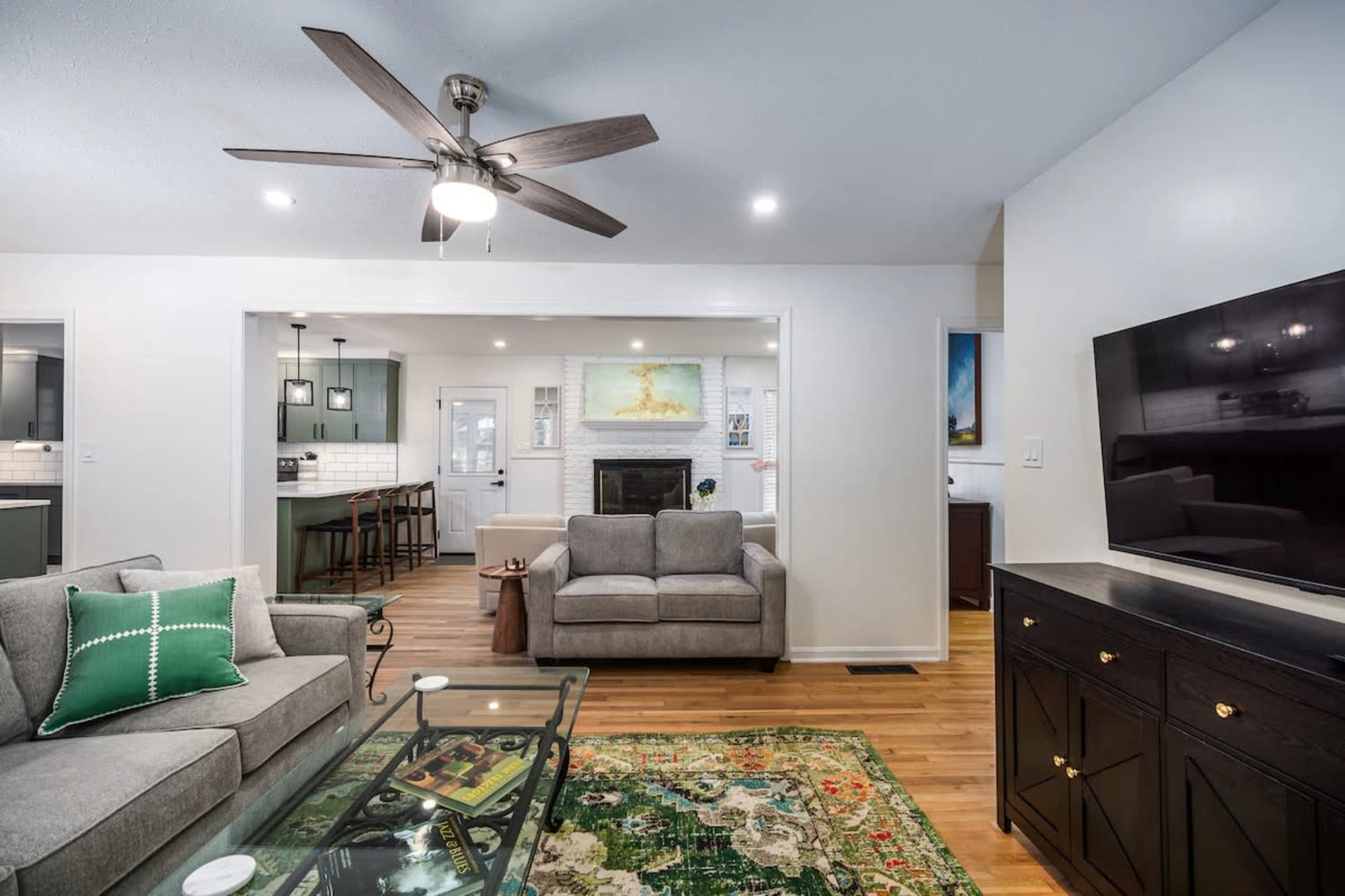 The image shows a modern living room with a gray sofa and armchair, a coffee table, a green area rug, and a flat-screen TV mounted on the wall, with a view into a connected dining area.