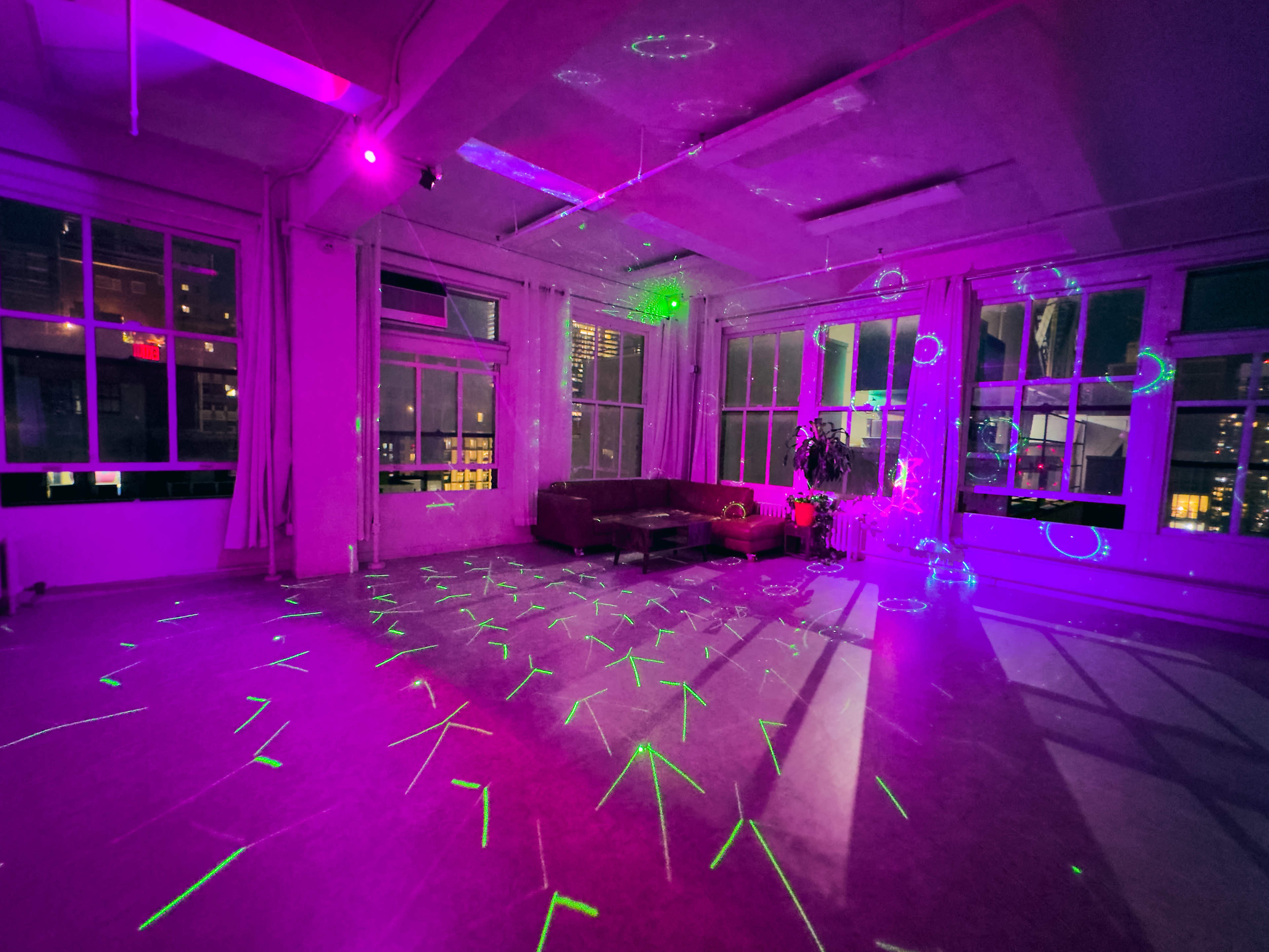 A brightly lit room features a couch and colorful laser patterns projected onto the floor, with large windows showing city lights outside.