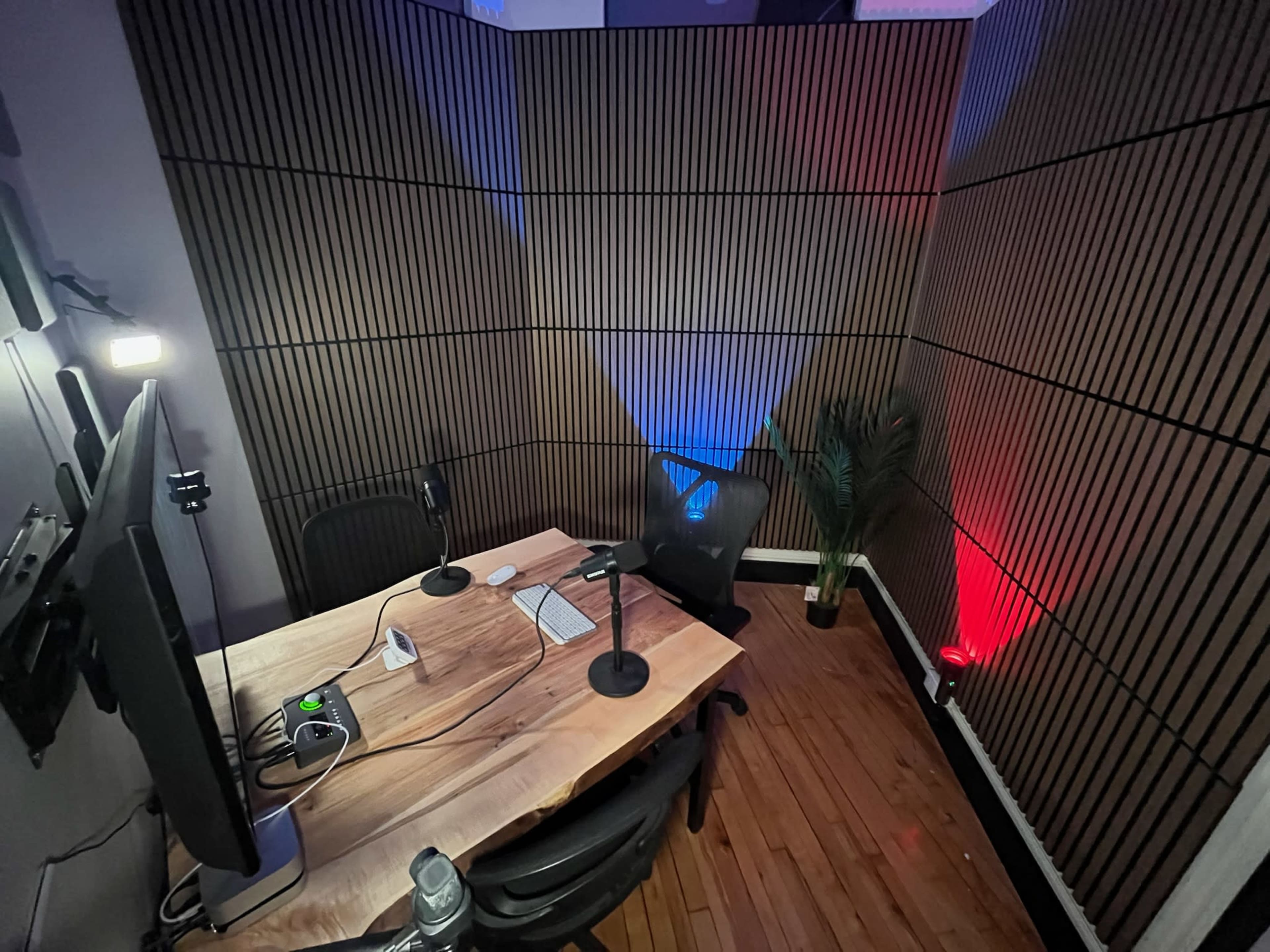 The image shows a dimly lit recording studio with a wooden table at the center, two microphones, a computer setup, and walls lined with wooden panels.