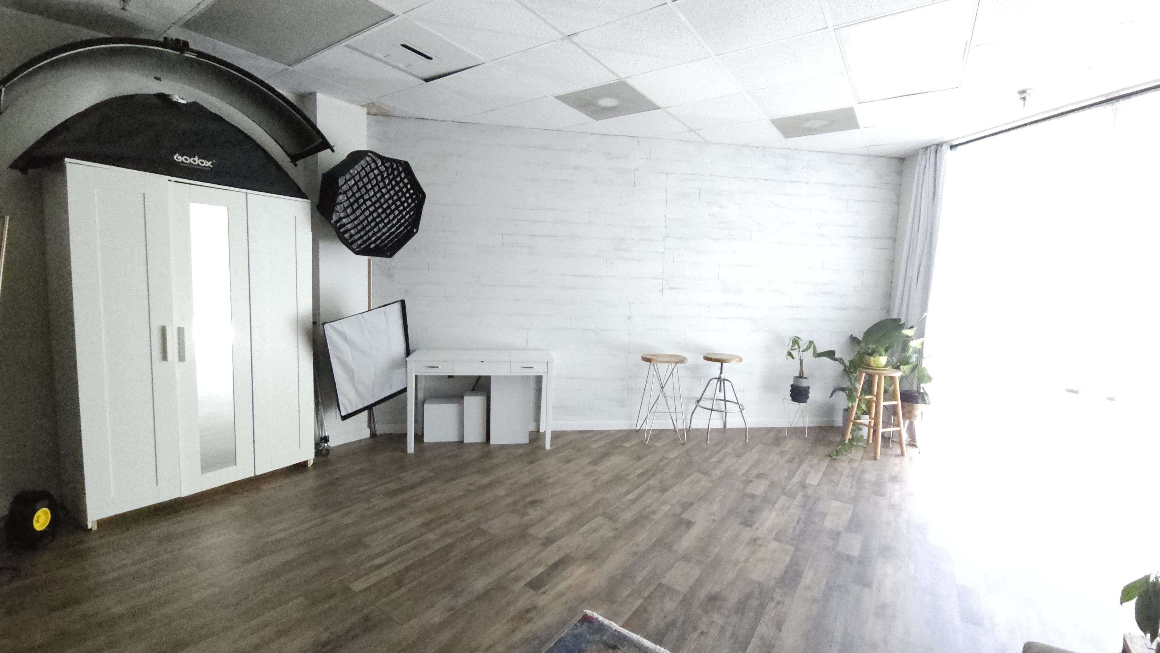 A spacious, minimalist studio with a light wooden floor, a white cabinet, a small desk, two stools, and large windows illuminating the area.