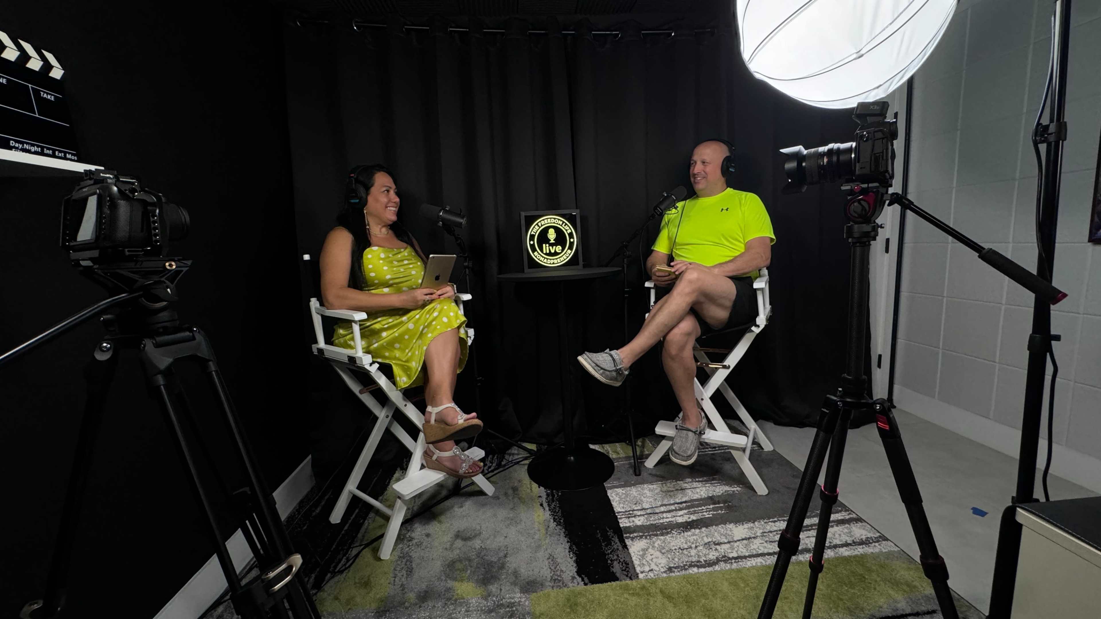 Two people sit in a recording studio, framed by black curtains, engaged in a conversation while microphones are positioned in front of them.