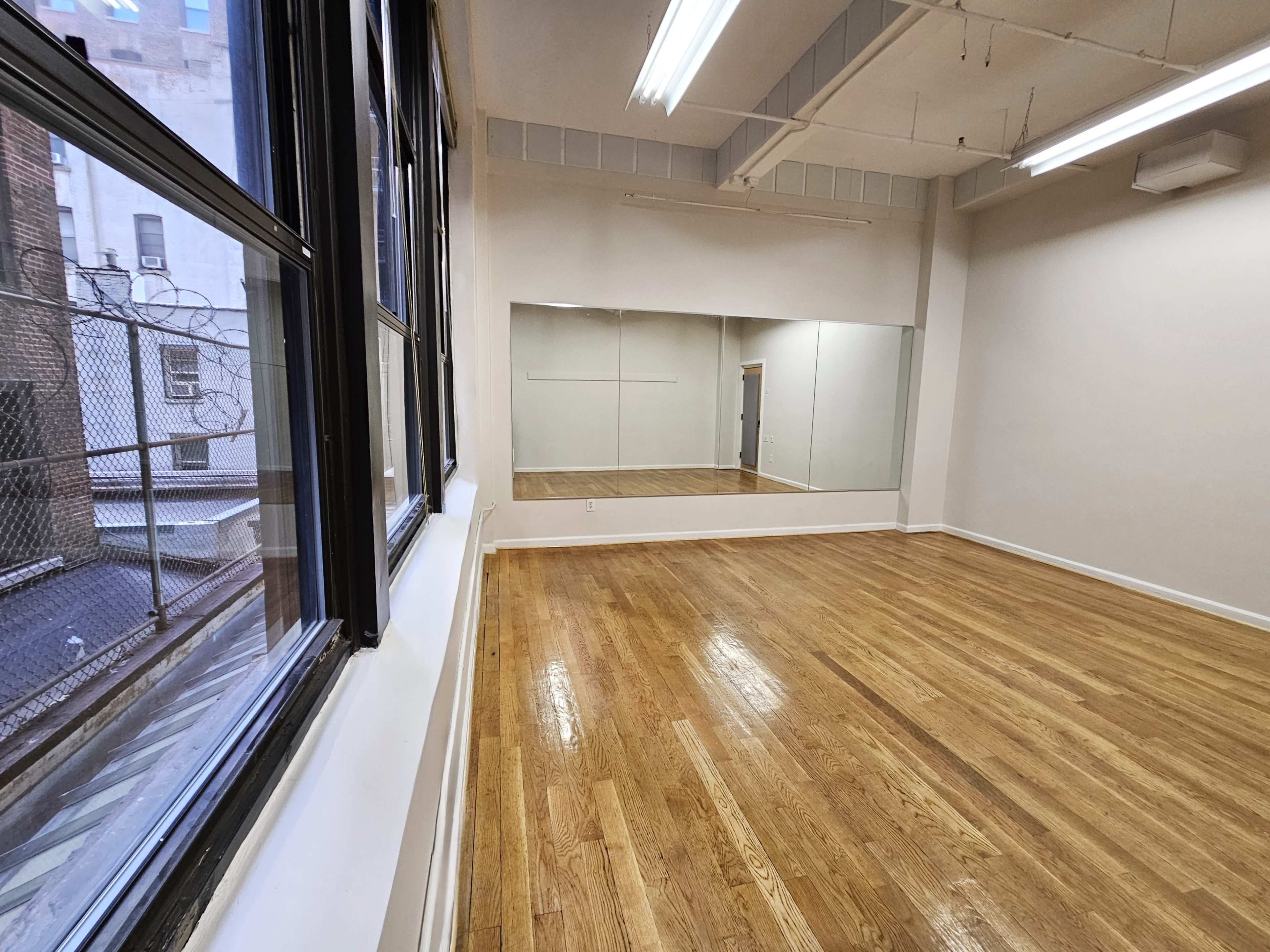 The image shows a spacious, empty room with hardwood floors, large windows, and a mirrored wall.