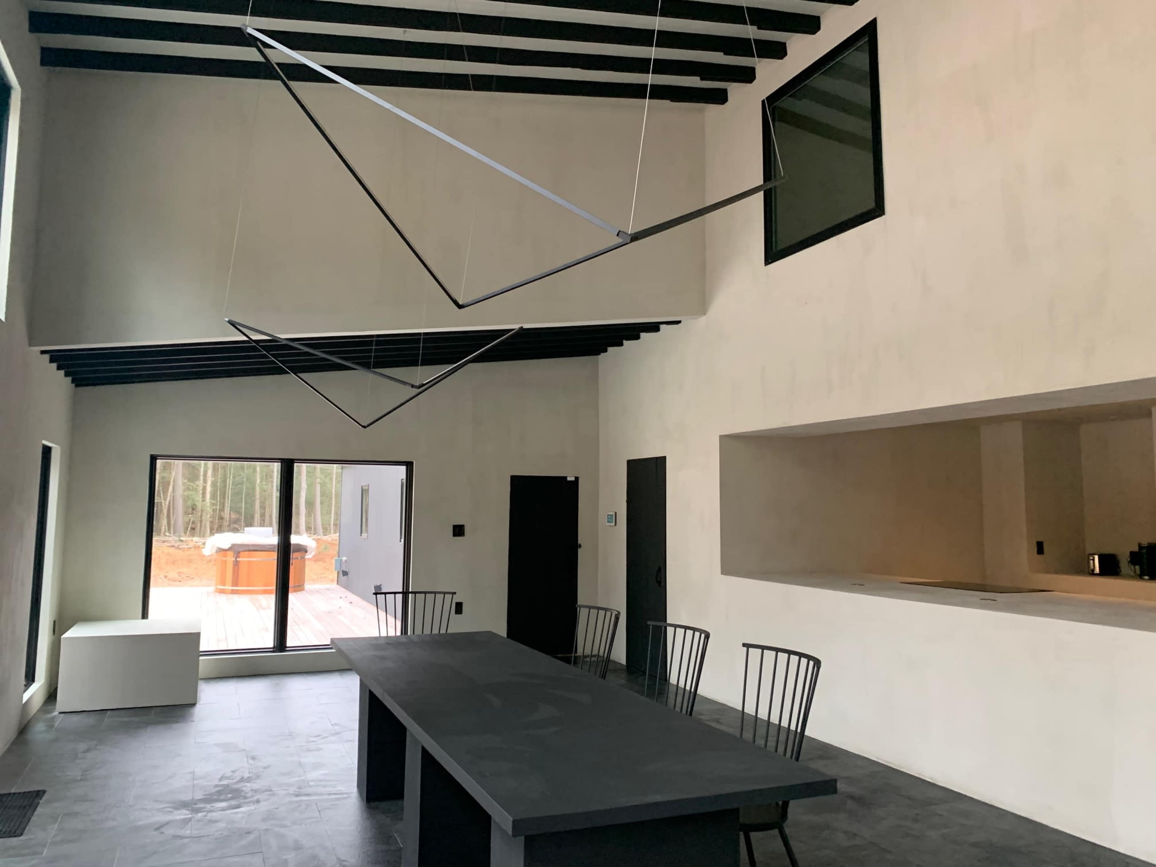 The image shows a modern interior space with high ceilings, featuring a long black table, four black chairs, and large windows that overlook a wooded area.