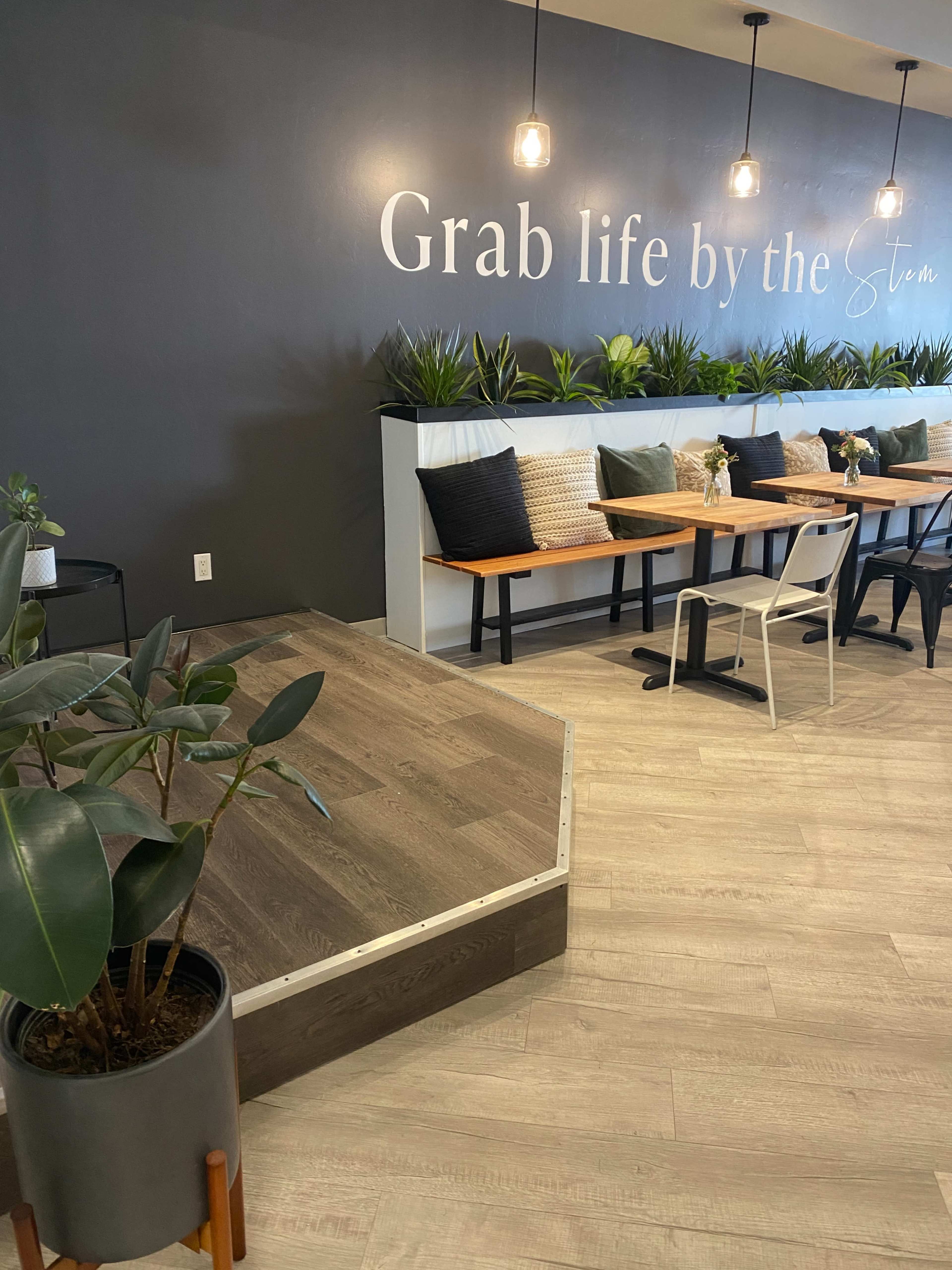 The image shows a cafe interior with wooden seating, decorative plants, and a wall that features the phrase "Grab life by the Stem."