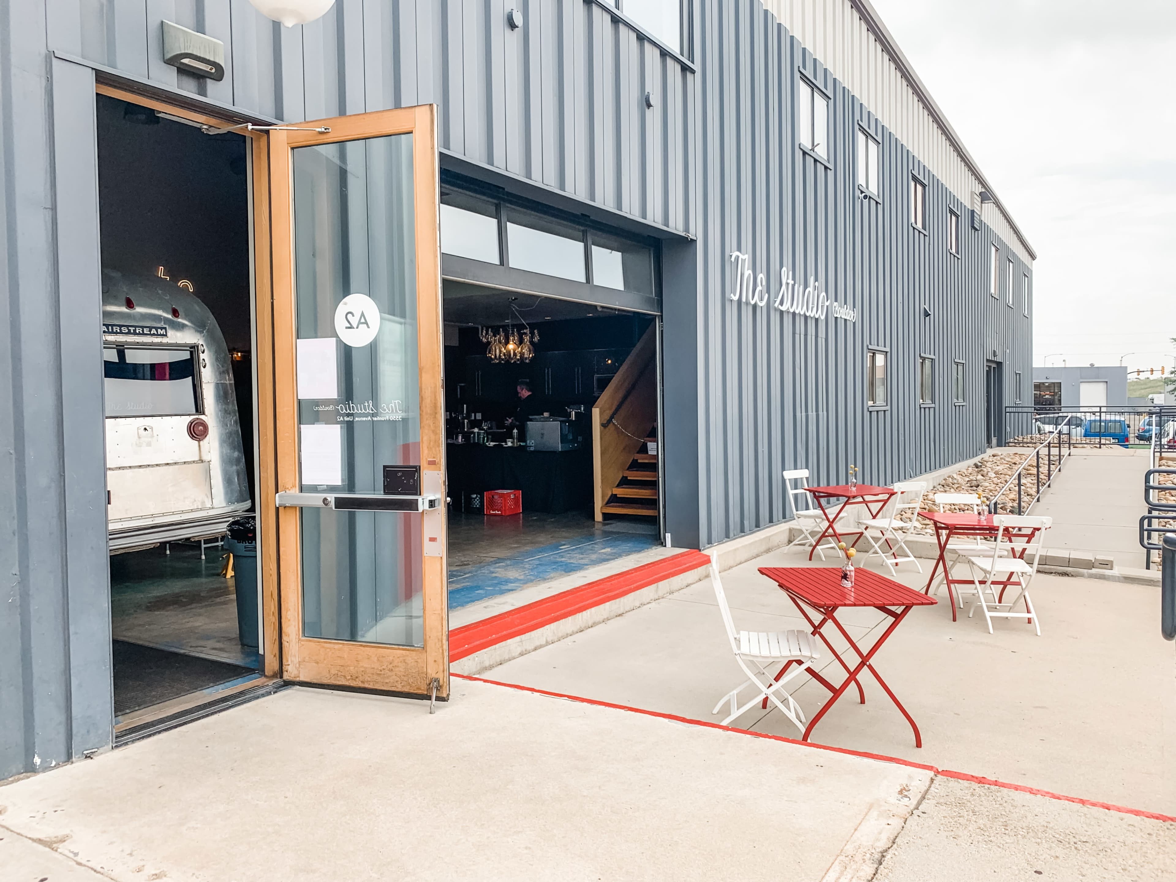 The image shows the entrance of a building with an open door, revealing an interior that includes stairs and a bar area, while two small red tables and chairs are placed outside on a concrete surface.