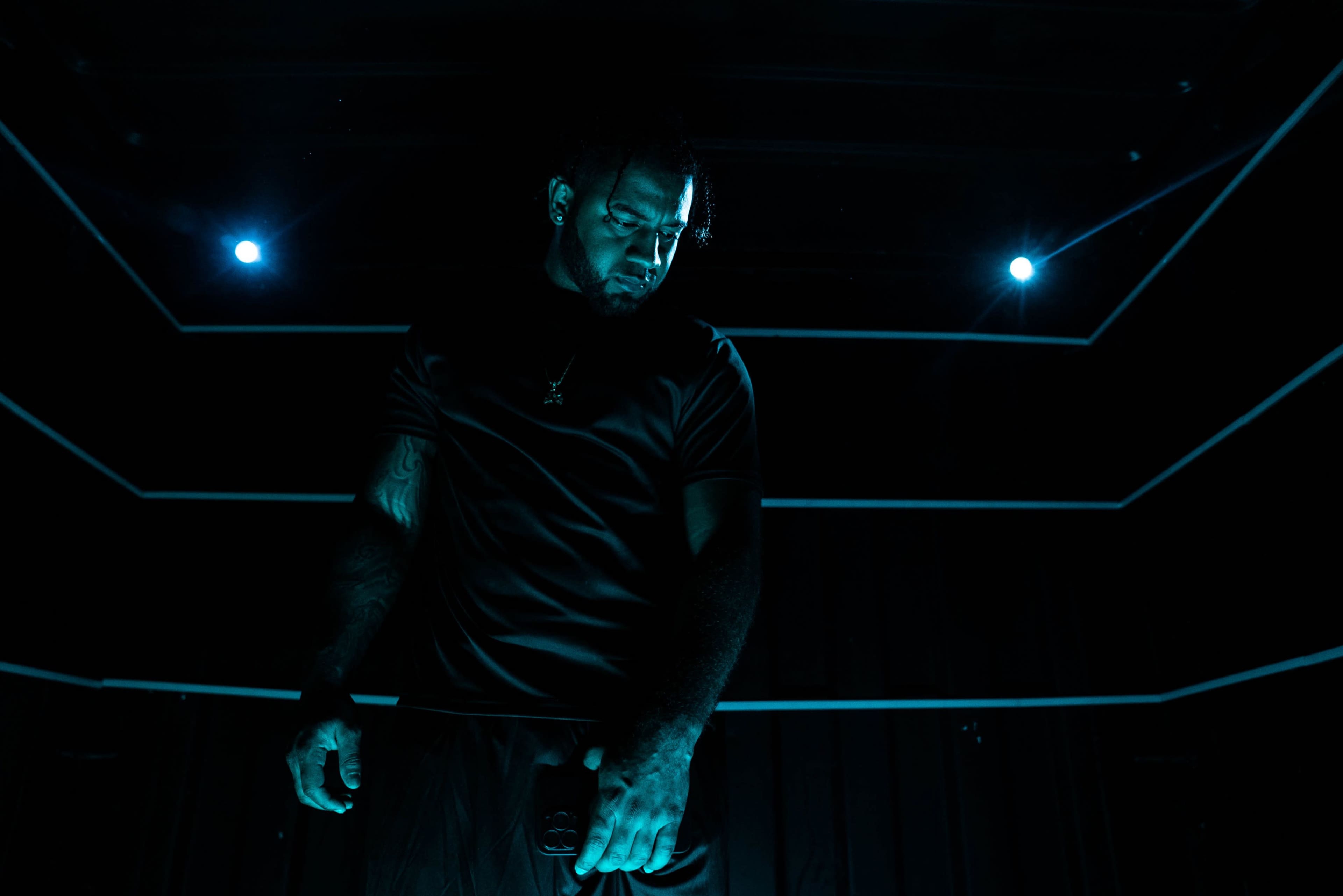 A man stands under bright blue lights, casting shadows against a dark background.