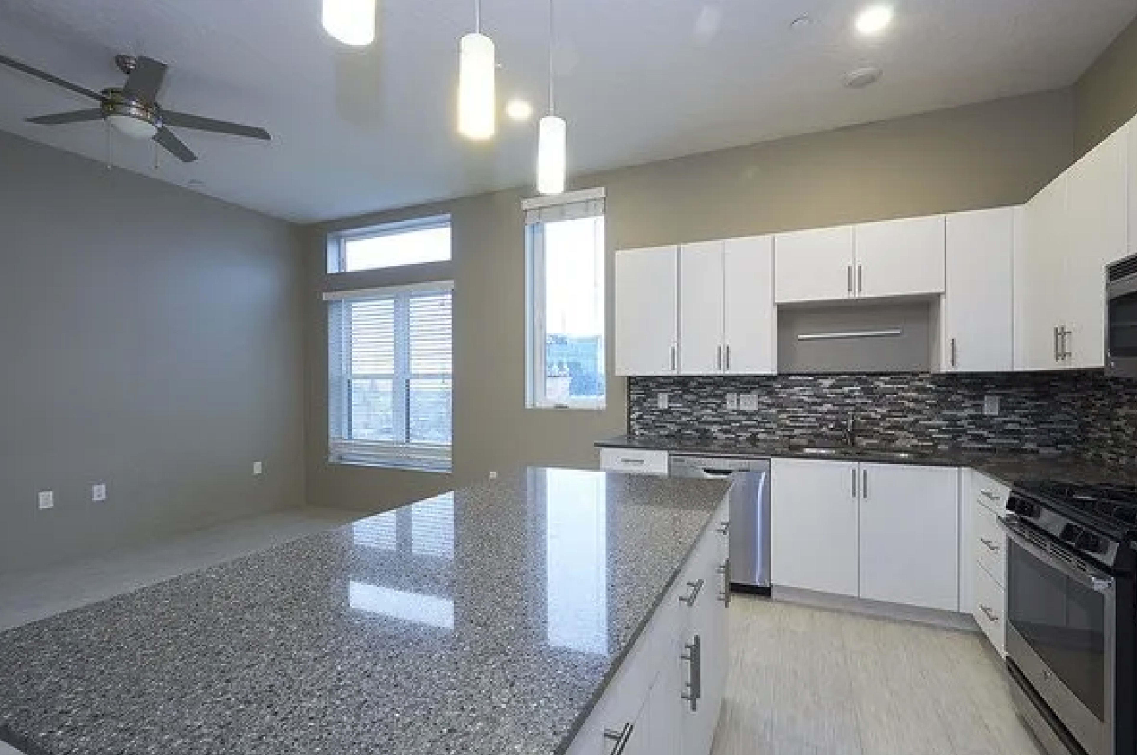 A modern kitchen with white cabinetry, a large granite countertop, and a tiled backsplash, complemented by pendant lighting.