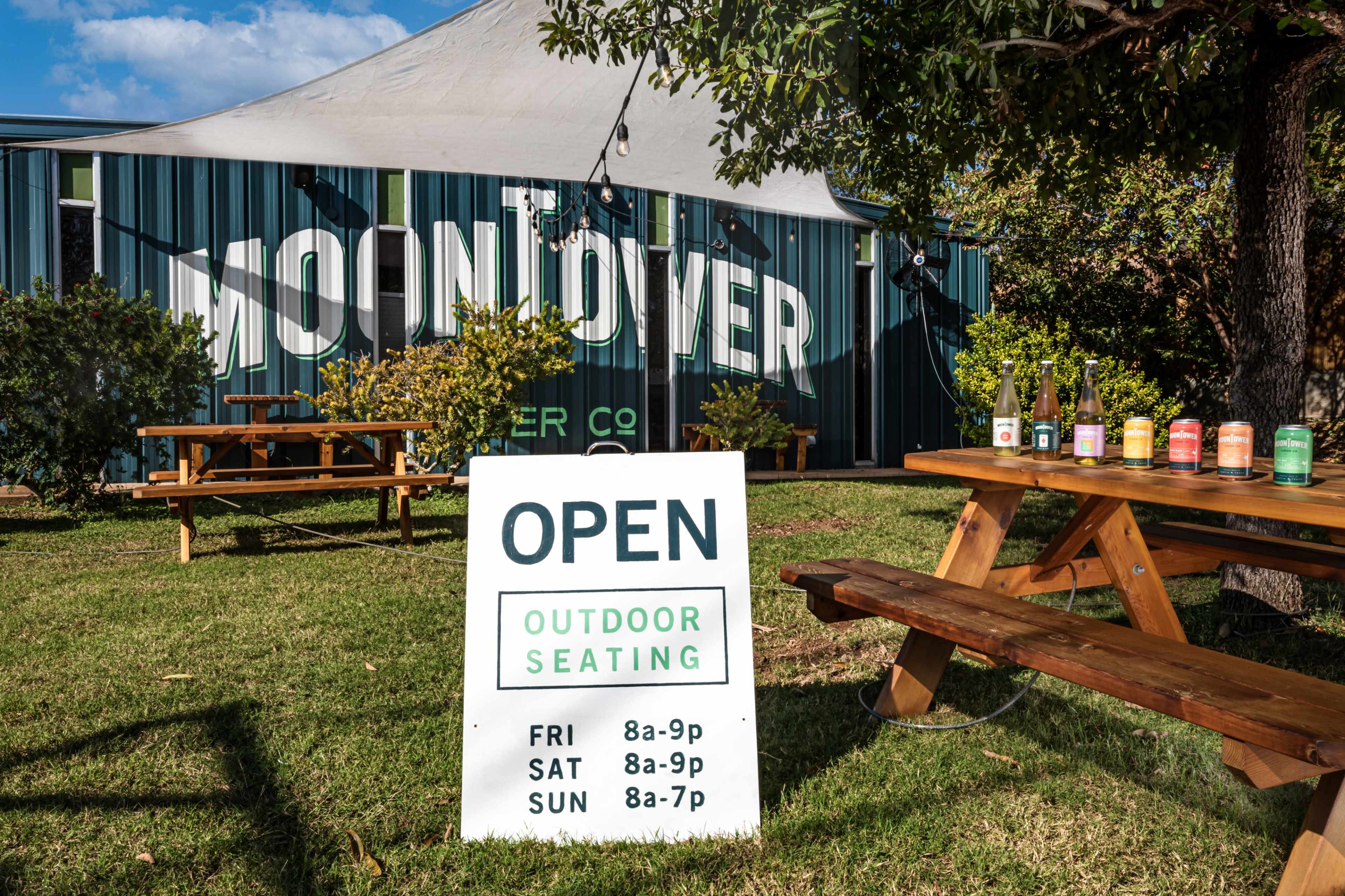 A sign indicating "Open" for outdoor seating, with a picnic table displaying drinks in front of a building labeled "Moon Over" in a green and white color scheme.