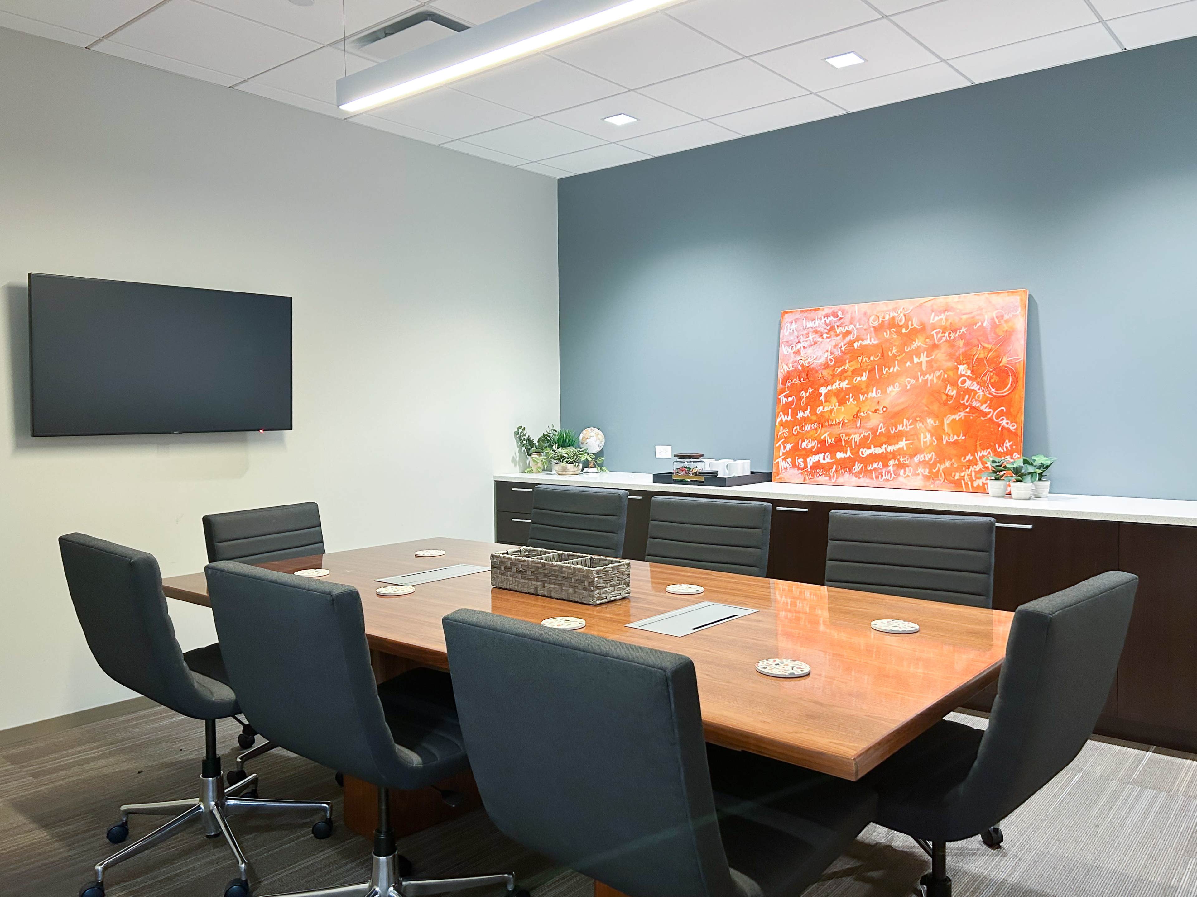 A modern conference room features a long wooden table surrounded by black chairs, a wall-mounted TV, and a colorful artwork hanging on the back wall.