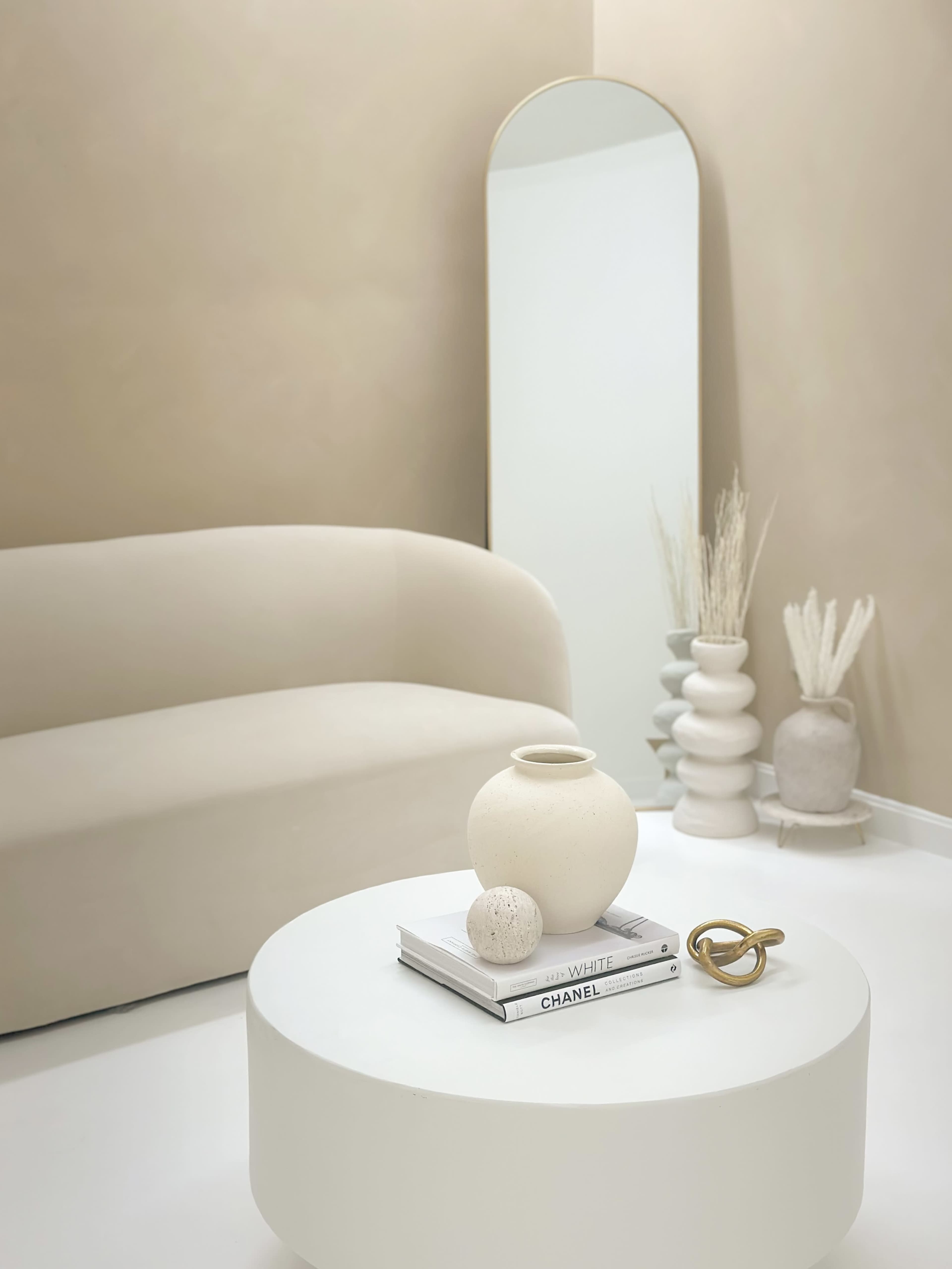 A minimalist living space featuring a curved beige sofa, a round white coffee table topped with a vase and a decorative orb, and a tall mirror against a neutral-colored wall.