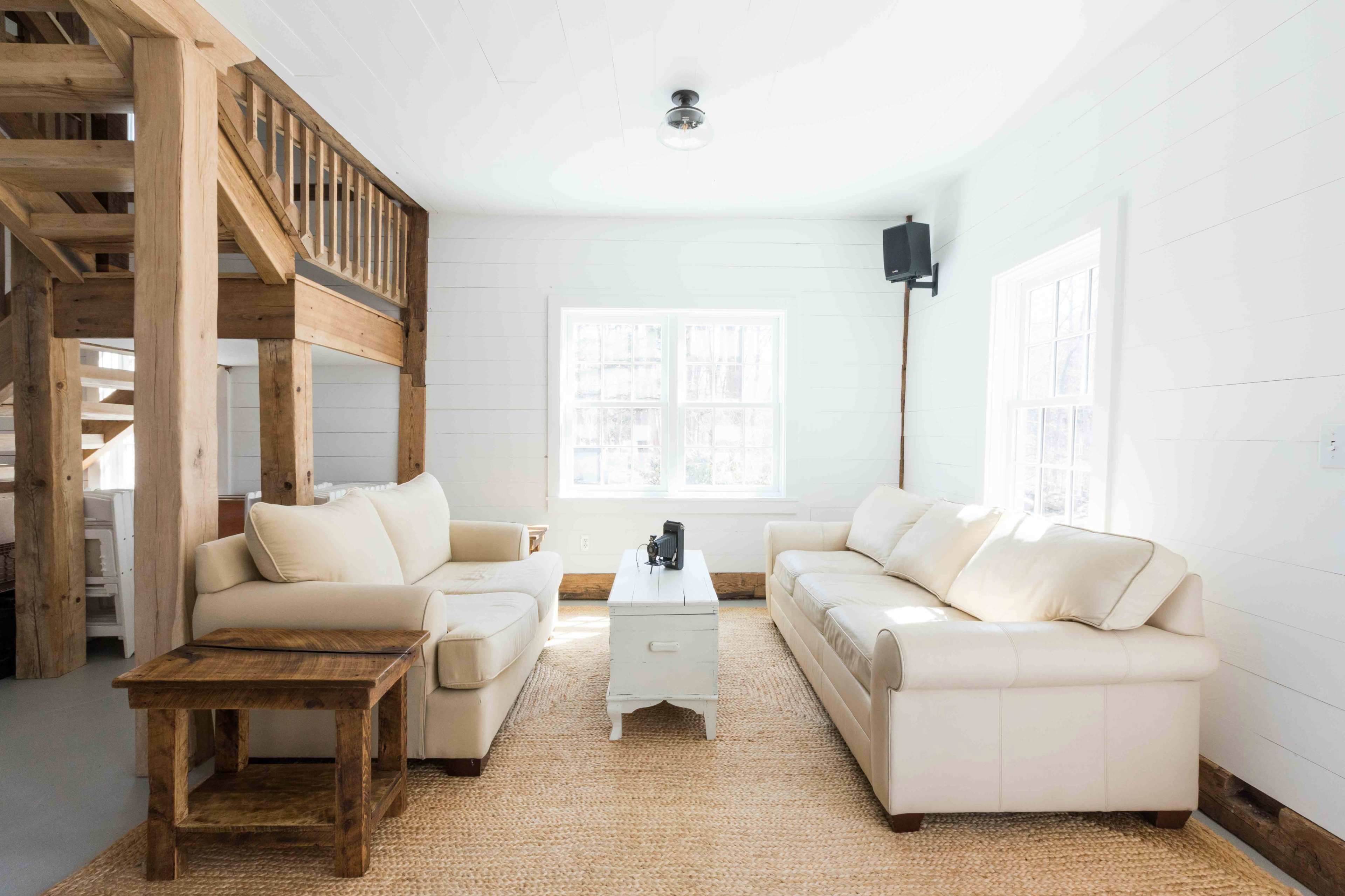 The living room features a light-colored leather sofa set arranged around a central white coffee table on a woven rug, with wooden accents and large windows letting in natural light.