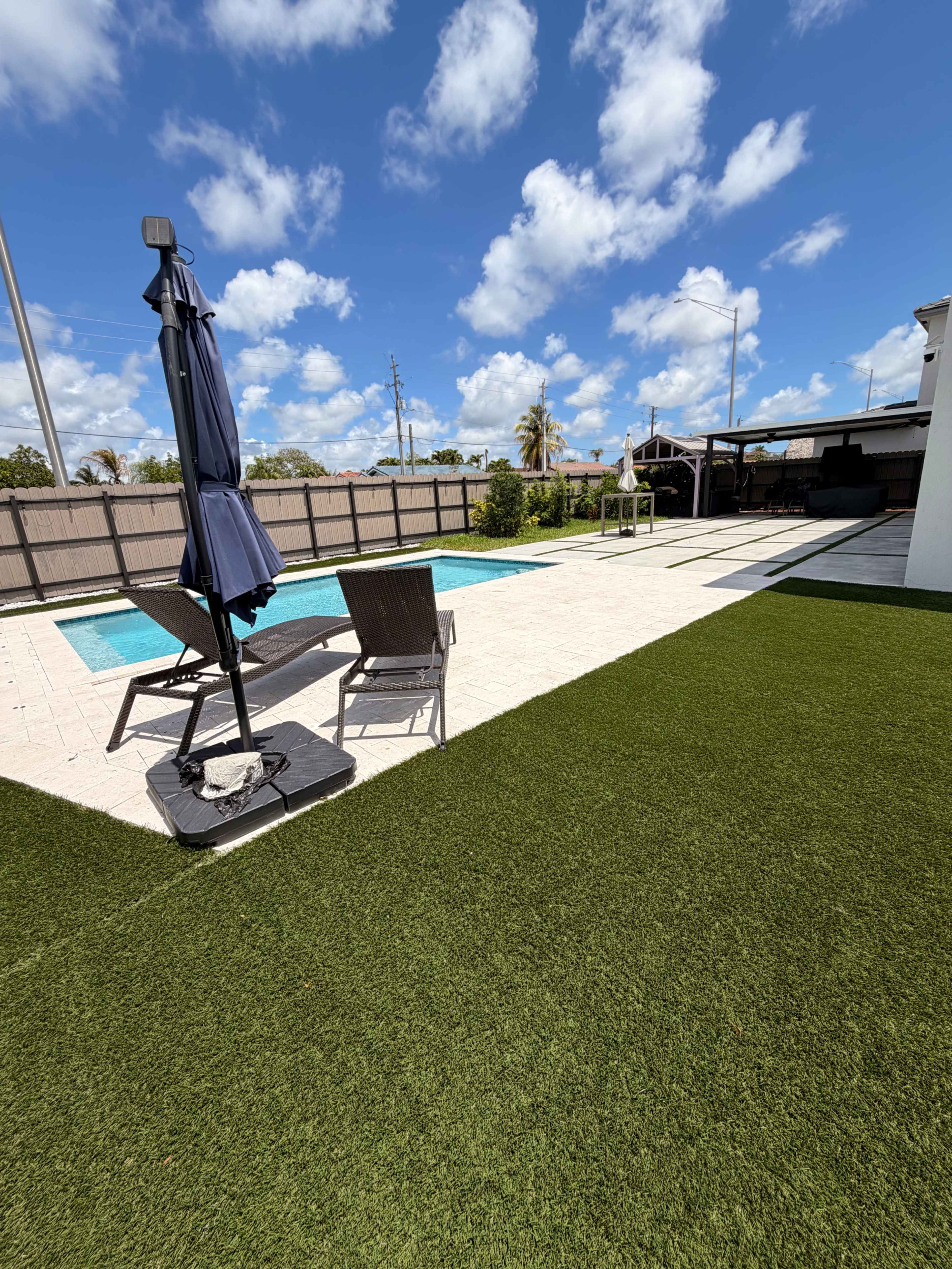 The image shows a backyard pool area with a swimming pool, two lounge chairs, a shade umbrella, and a grassy lawn under a blue sky with clouds.