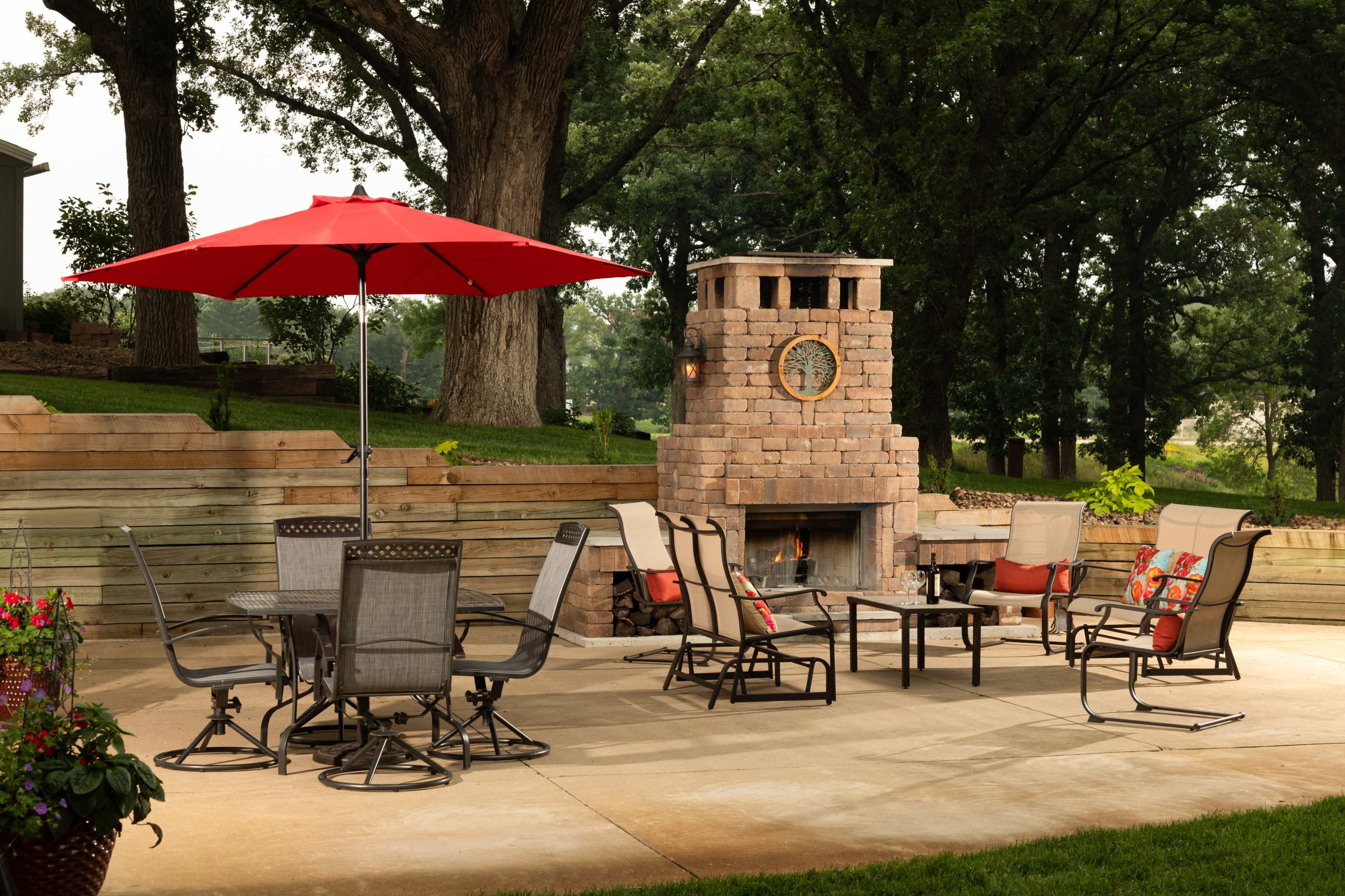A stone outdoor fireplace is surrounded by a seating area featuring metal chairs and a table, under a red umbrella on a concrete patio.