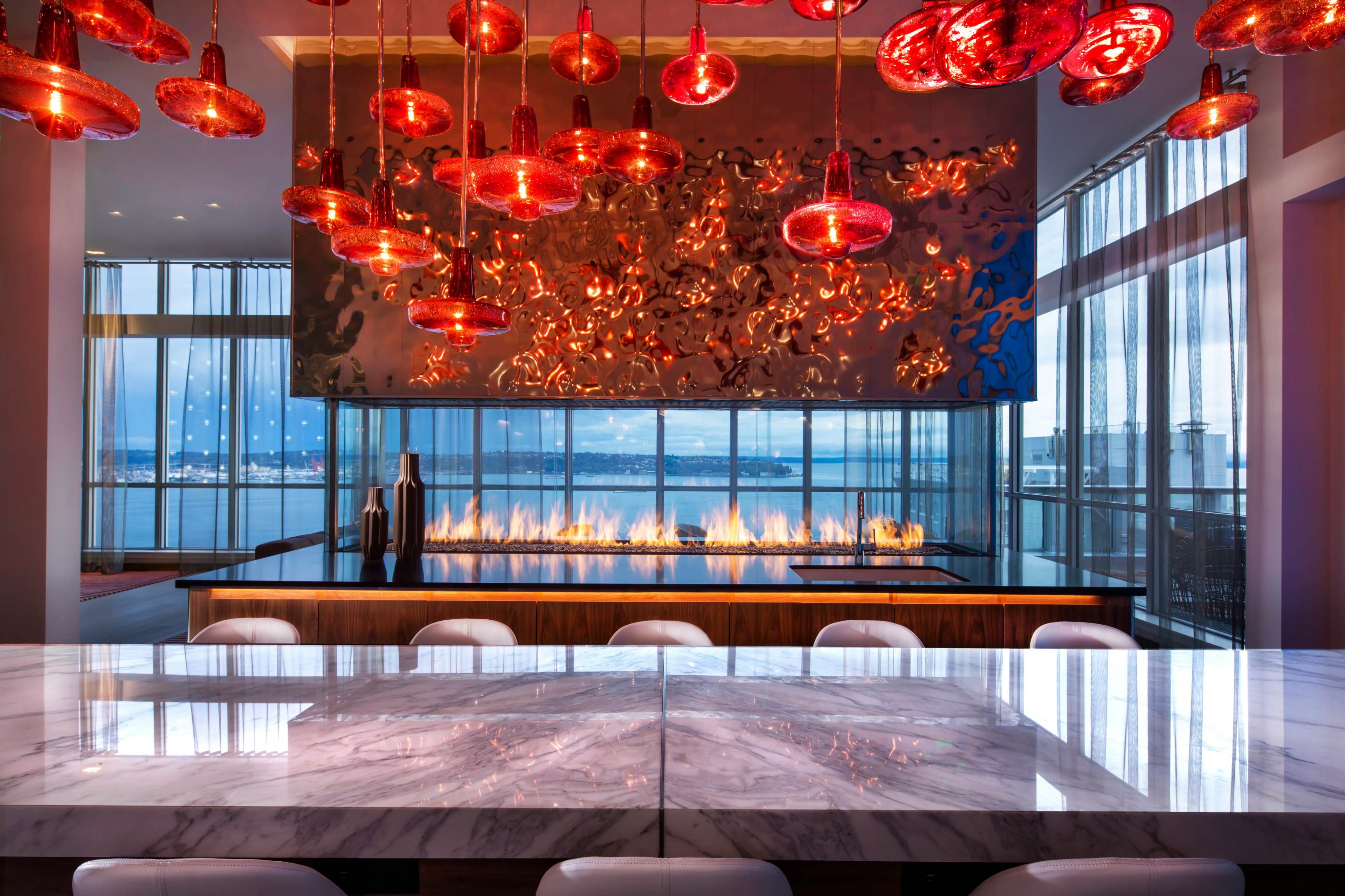 A luxurious dining area features a large marble table, an open kitchen with a modern design, and striking red glass pendant lights above, with a view of water and a fireplace in the background.