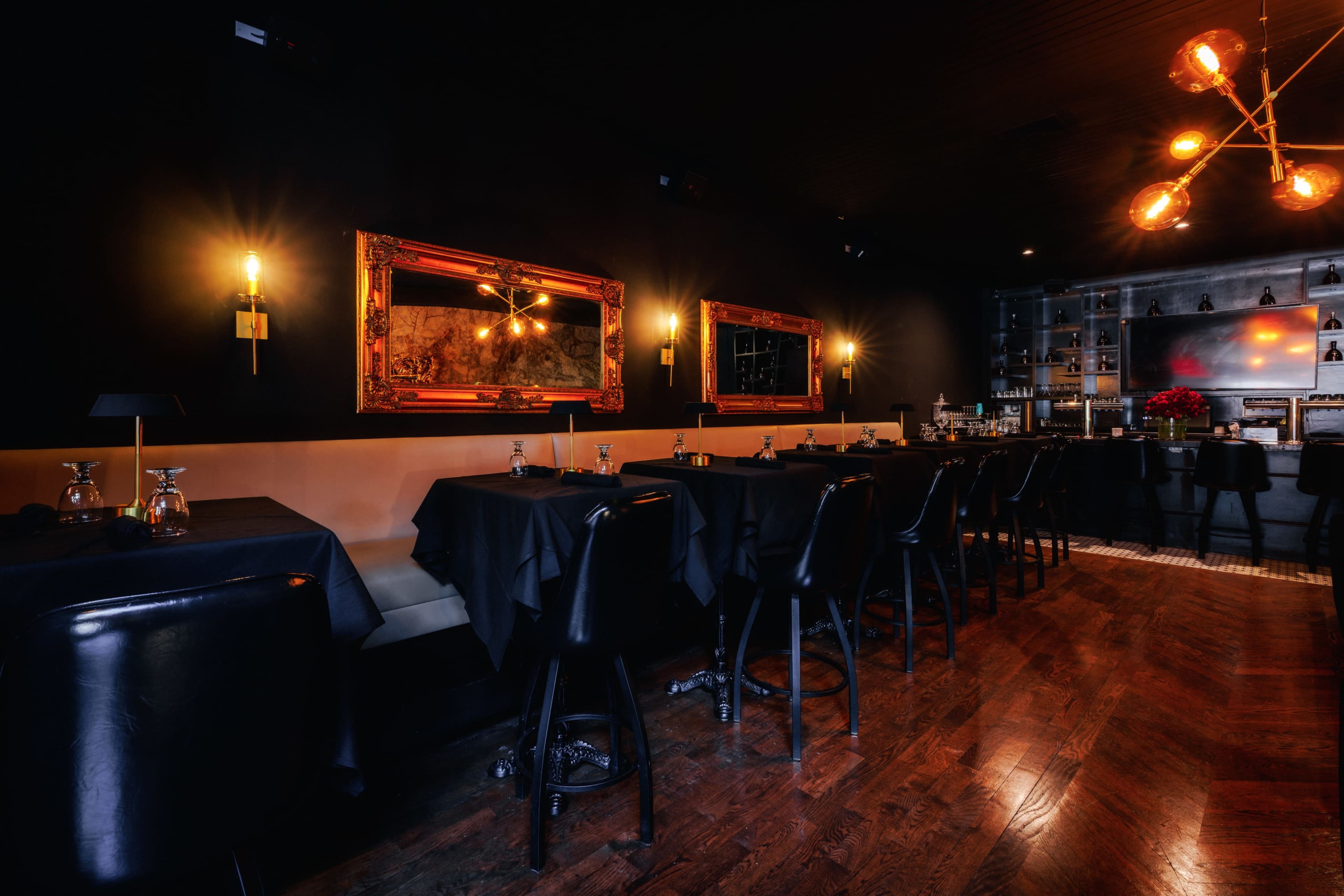 The image shows a dimly lit restaurant interior featuring black walls, dark wooden flooring, and tables set with black tablecloths.