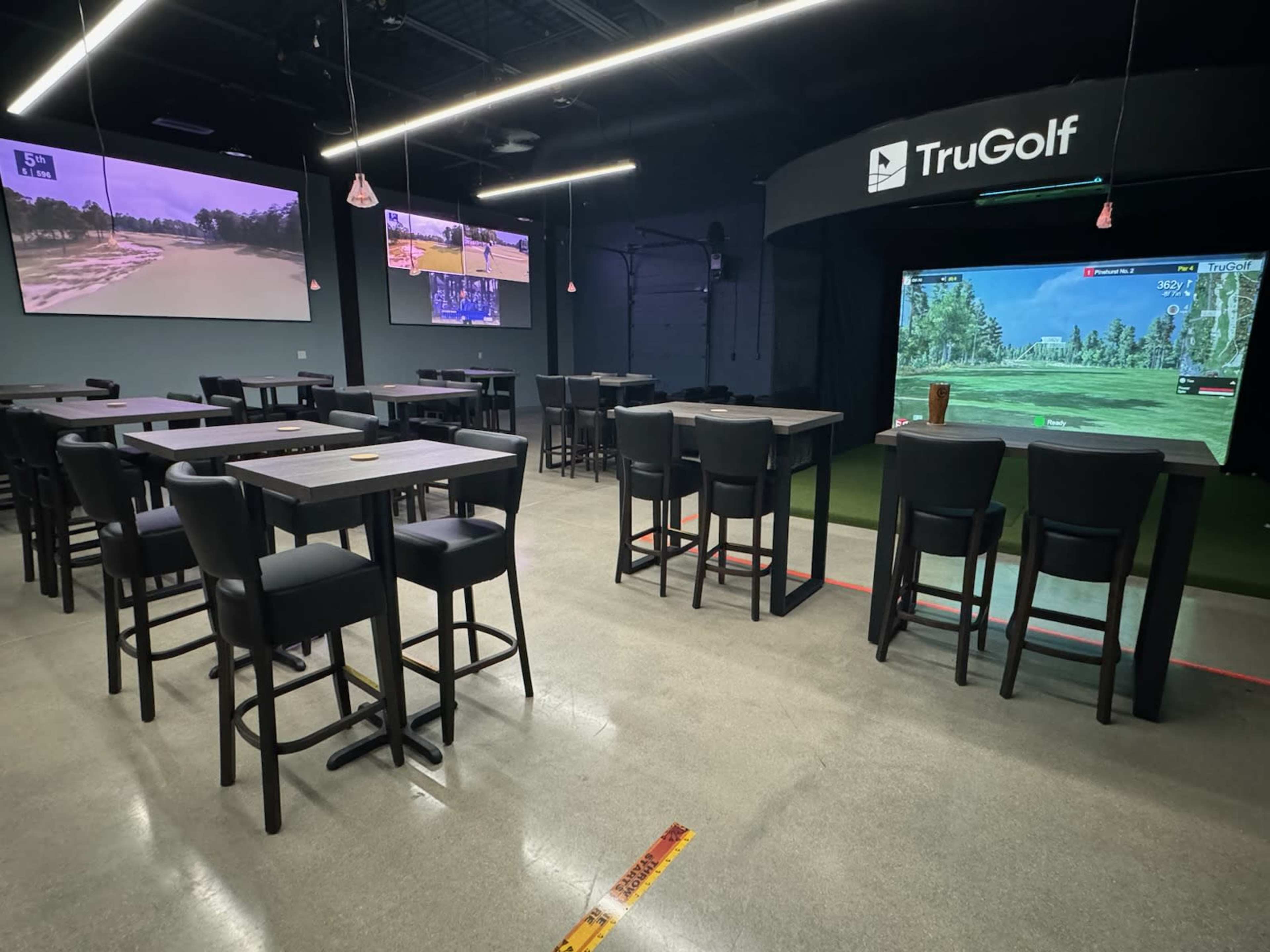 An indoor golf simulation facility with multiple screens displaying golf courses, surrounded by high tables and bar stools.