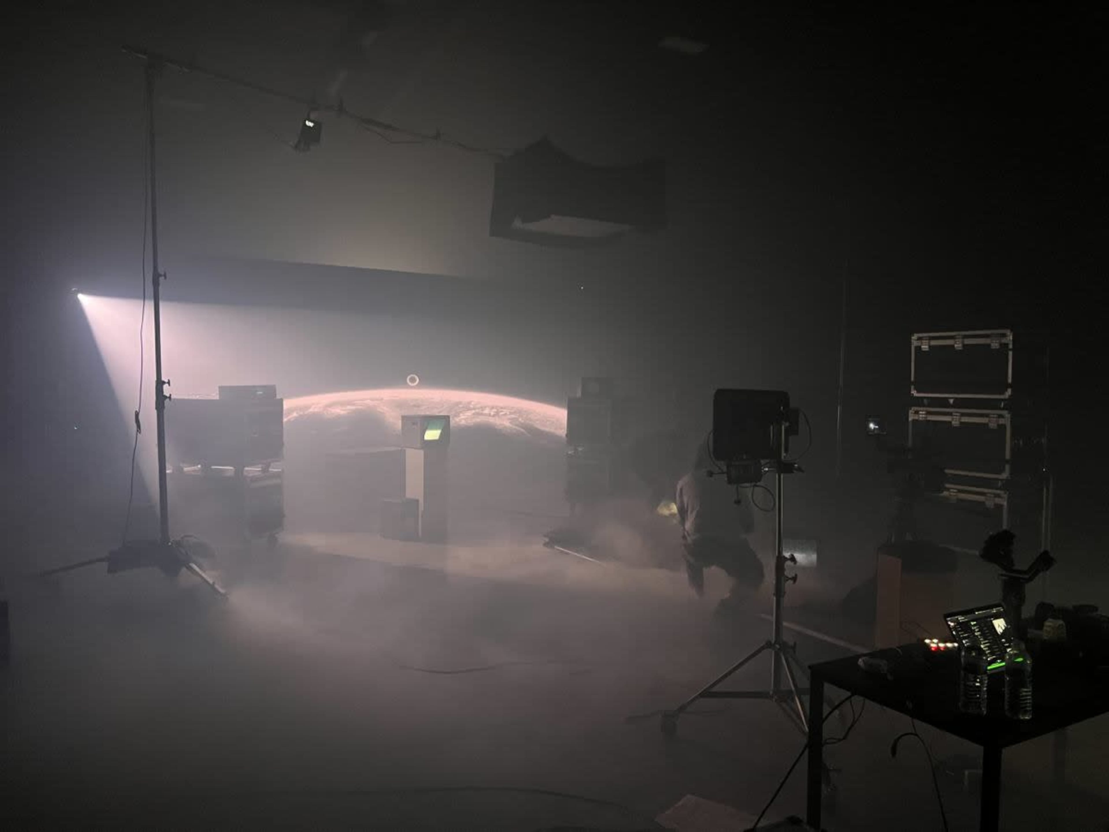 A dimly lit studio setup features a monitor displaying a planetary image, surrounded by fog and various equipment.