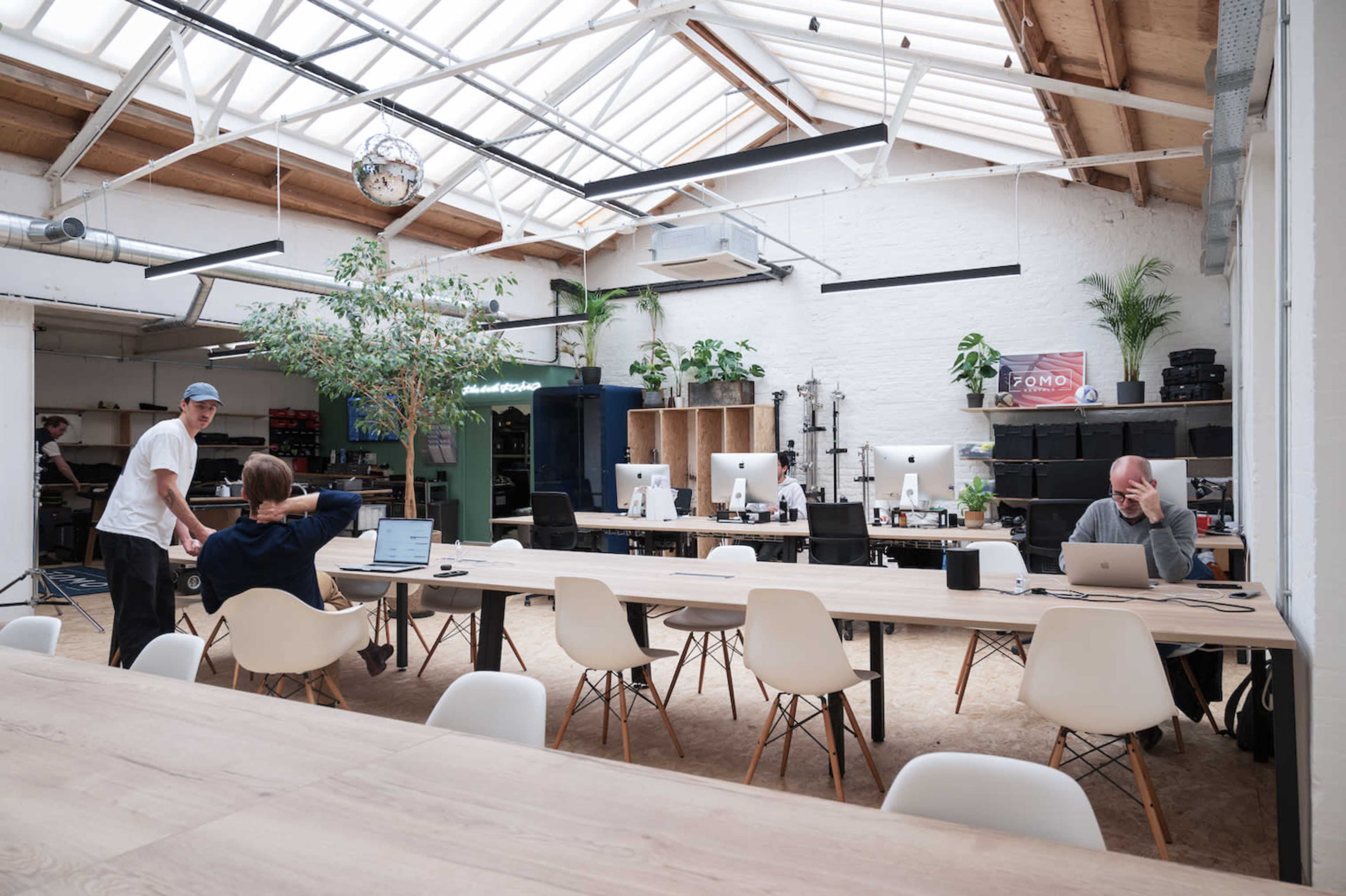 A spacious coworking space features several desks with computers, a large communal table, and plants, all under a glass ceiling.