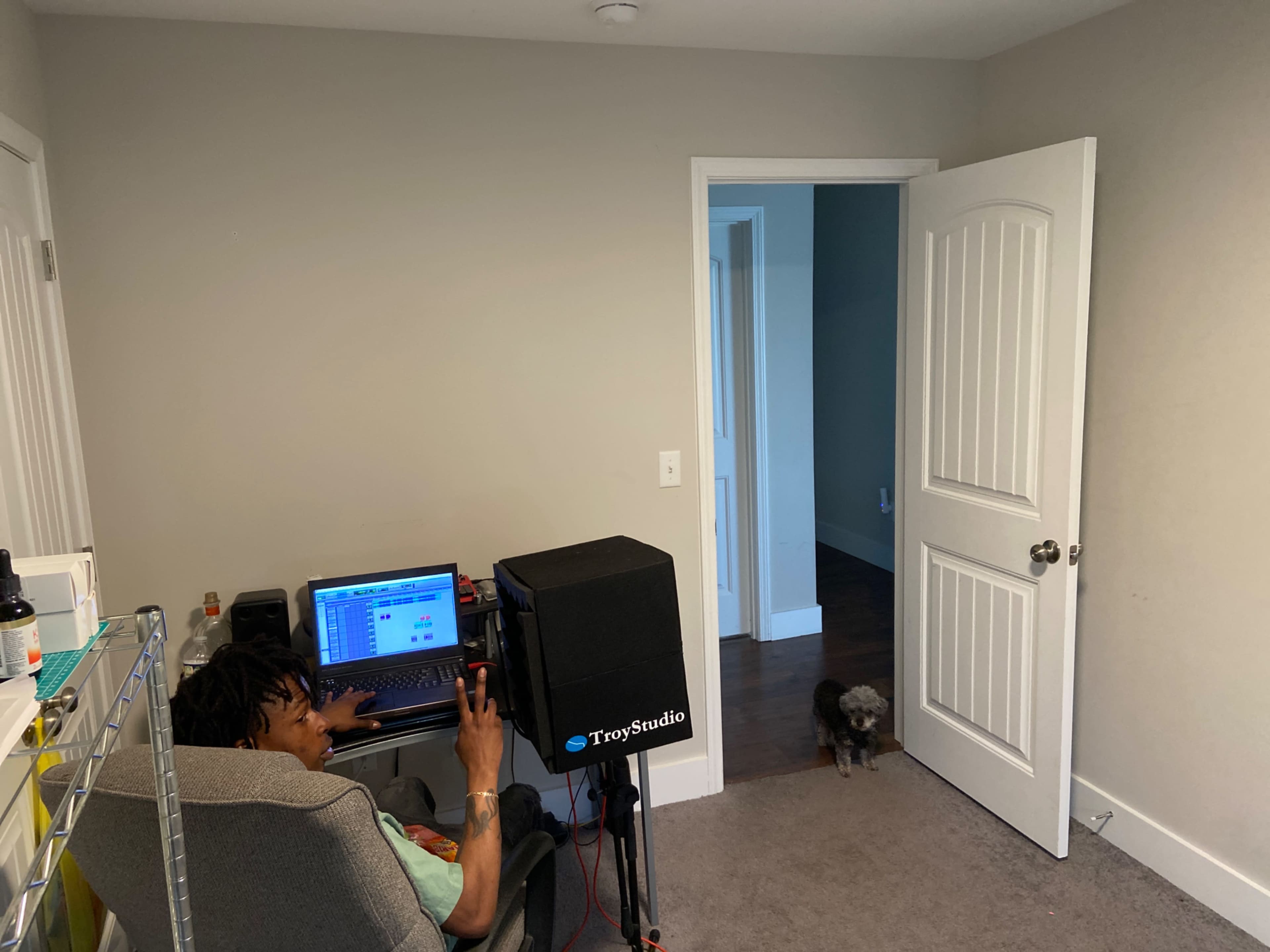 A person sits at a desk with a laptop and a microphone in a sparsely furnished room while a dog walks near an open door.