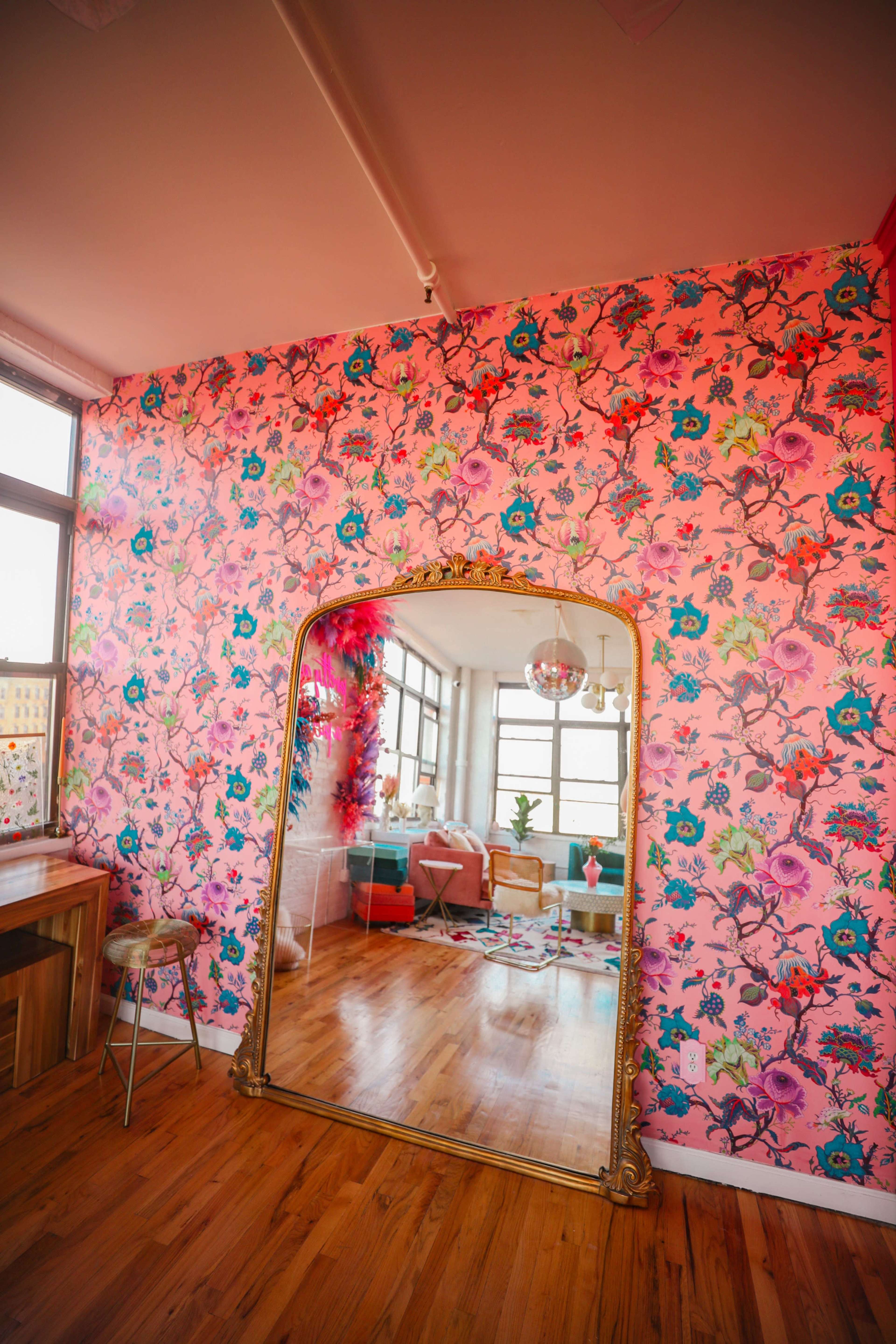 The room features a large ornate mirror reflecting a brightly patterned floral wallpaper and a living area with a green sofa and potted plants.