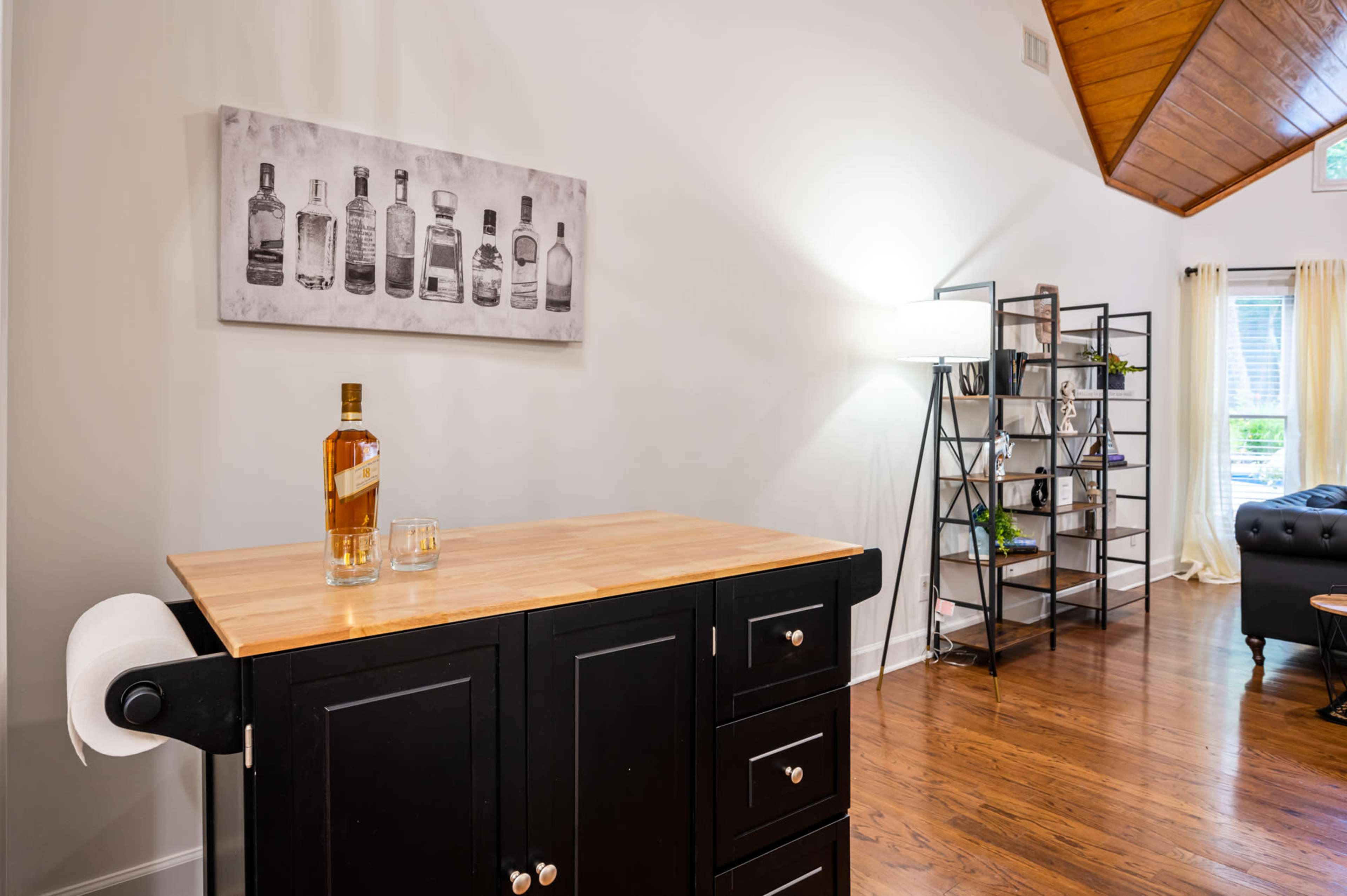 A wooden kitchen cart with a bottle of liquor and a glass sits next to a tall bookshelf in a well-lit room featuring a sloped ceiling.