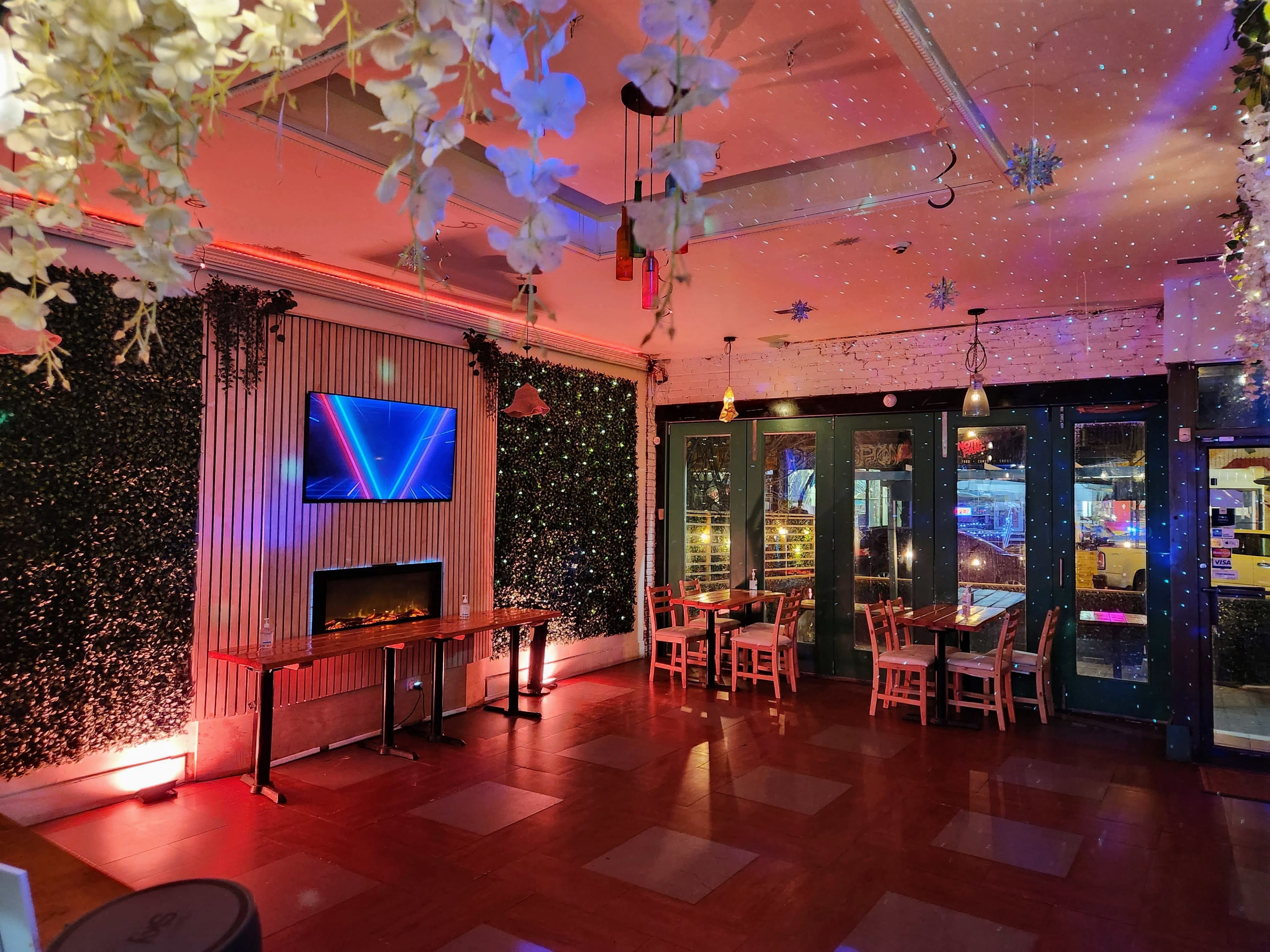 The interior of a restaurant features green foliage on the walls, a lit-up fireplace, and a large screen displaying vibrant graphics, with tables and chairs arranged near windows showing city views.