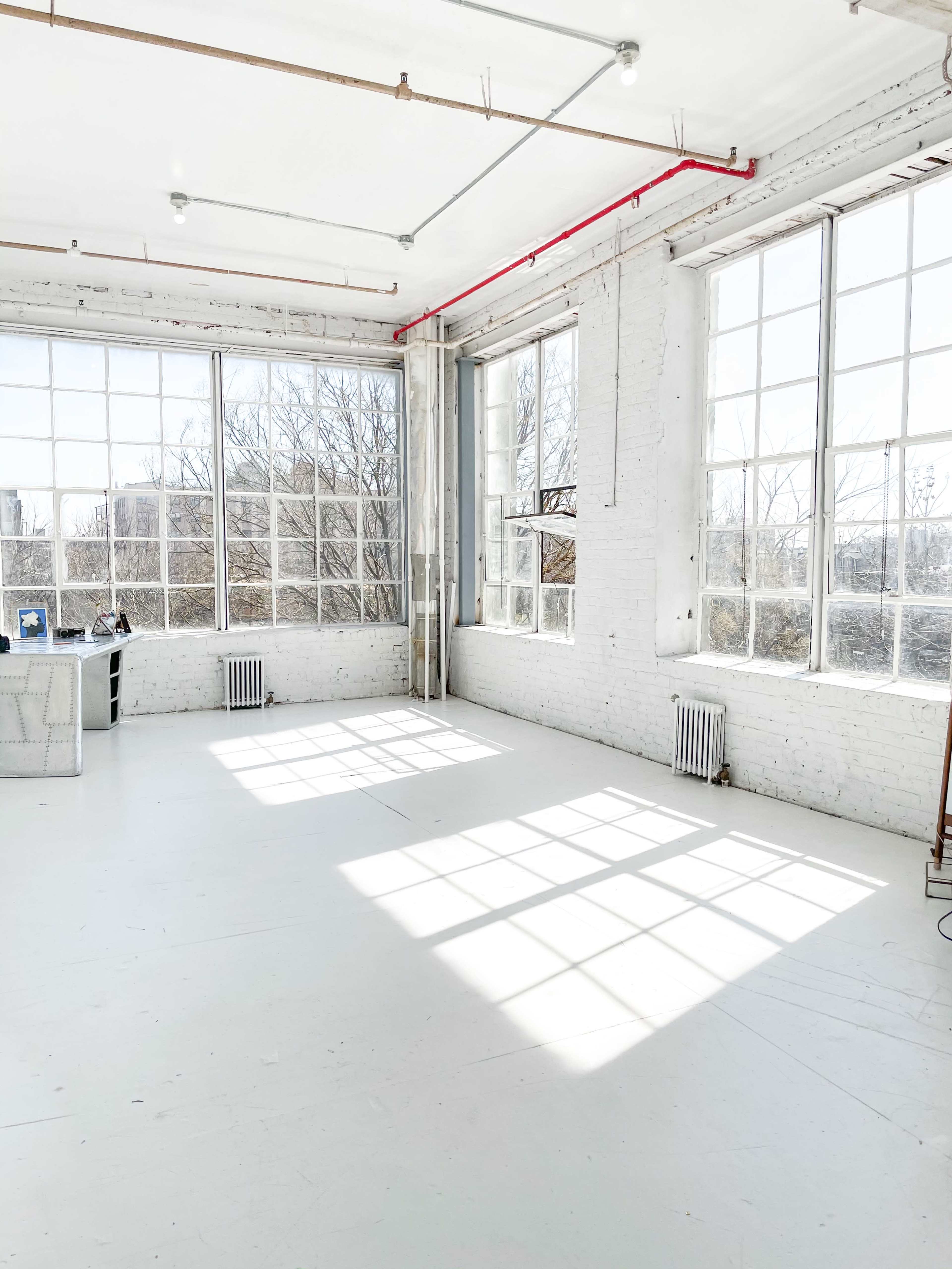 A bright, empty room with large windows and white walls, featuring visible shadows on the floor.