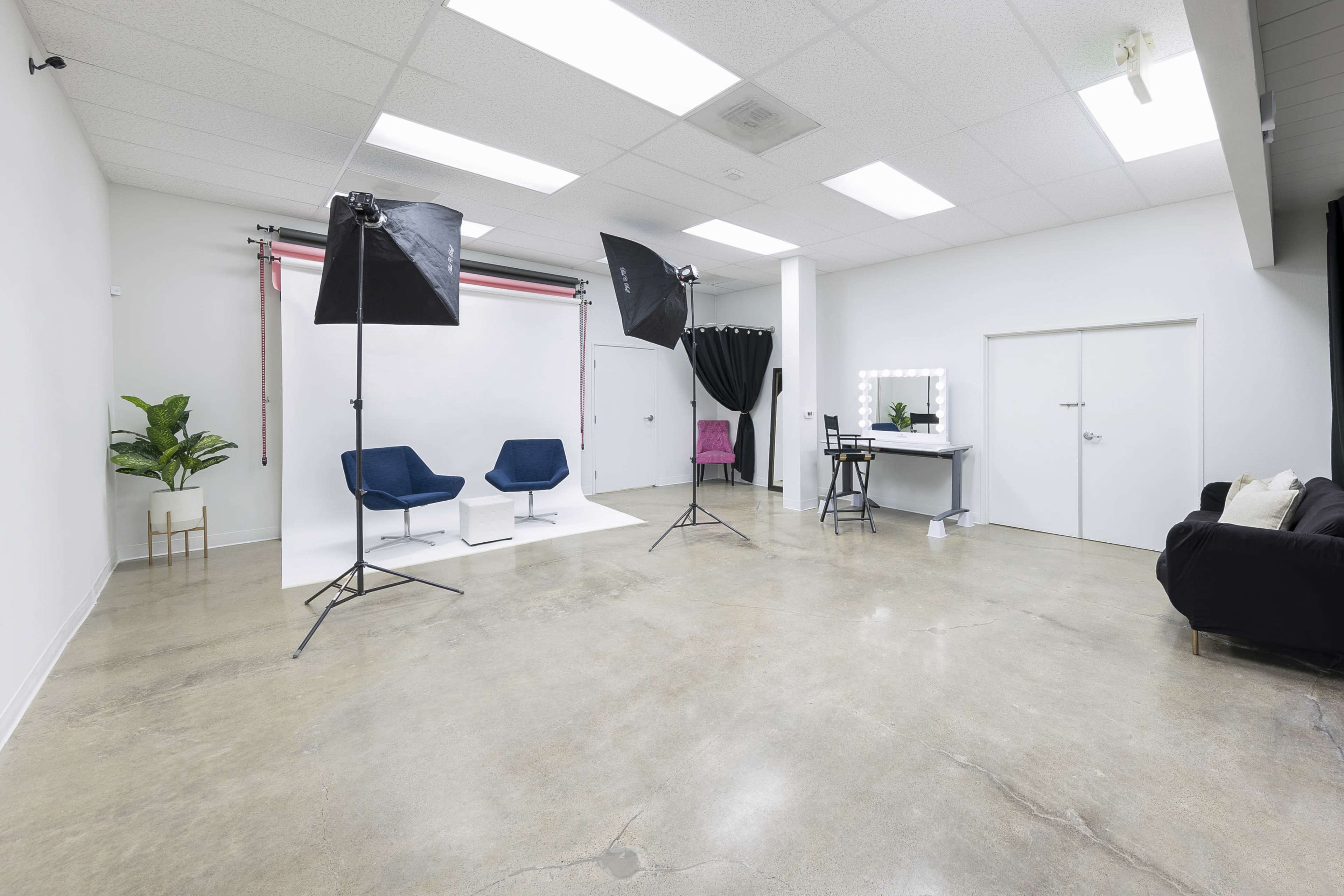 The image shows a photography studio with two blue chairs, two large softbox lights, and a makeup station against a plain white backdrop.
