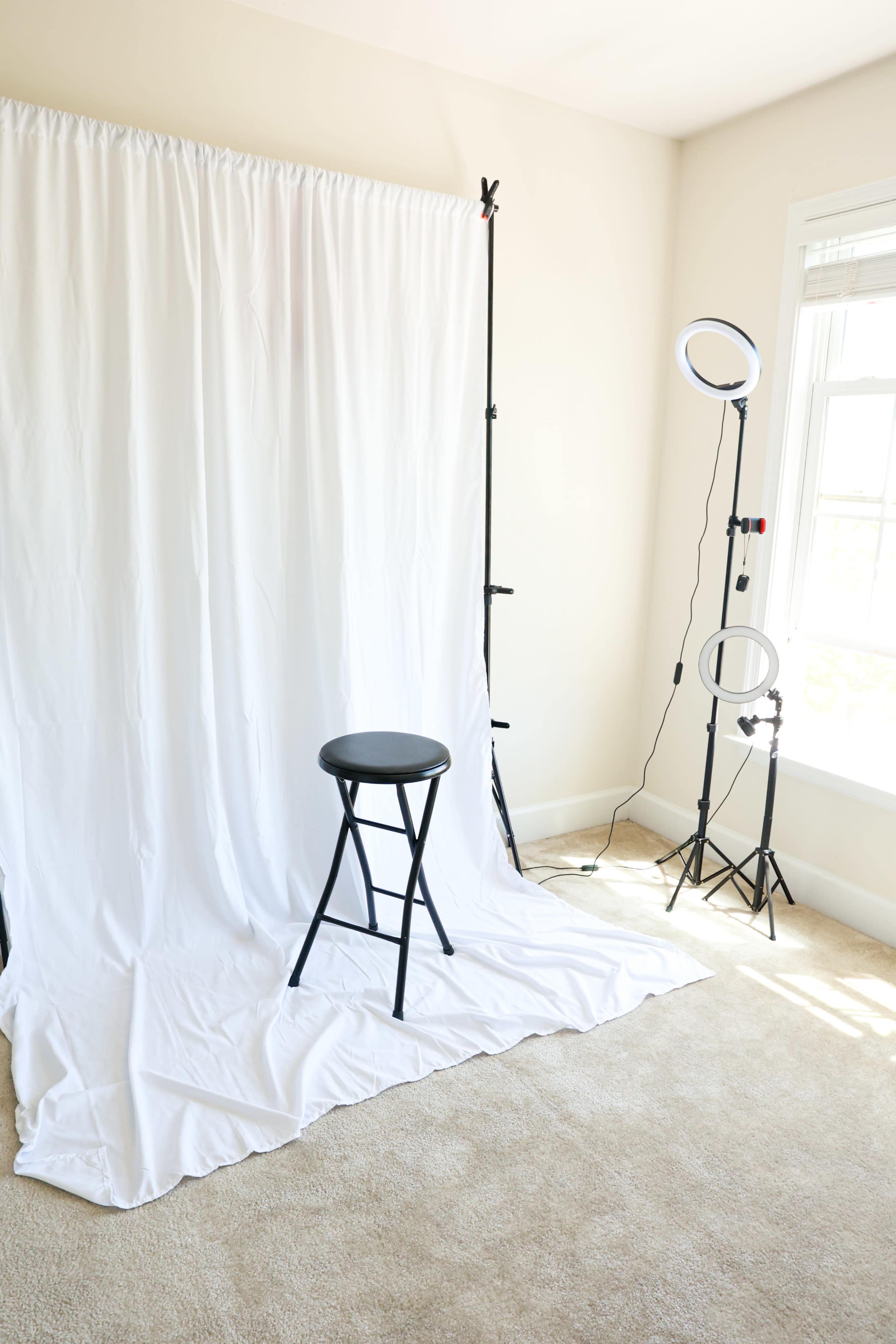 The image shows a spacious room with a white backdrop, a black stool in the center, and lighting equipment positioned to the side.