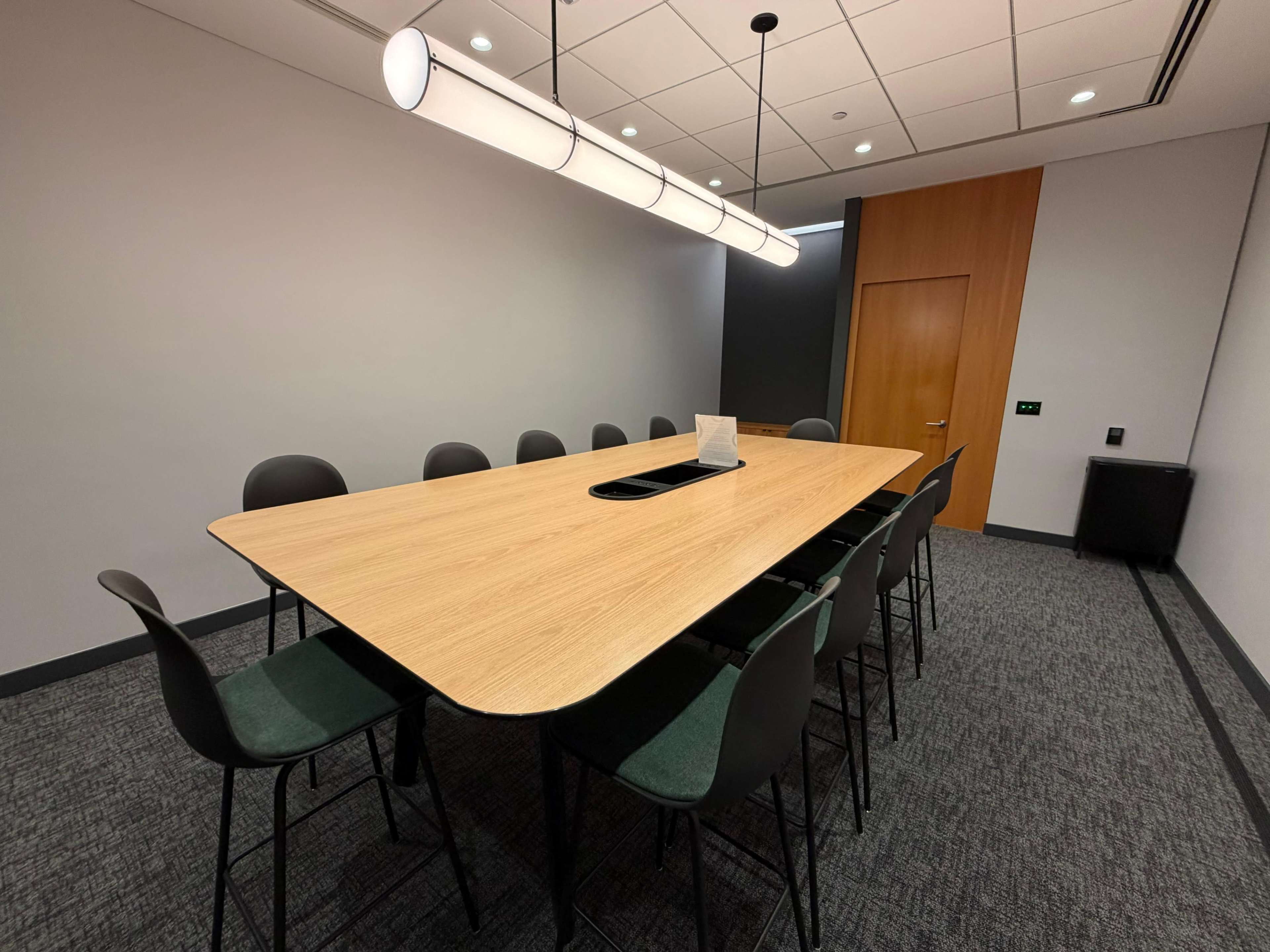 The image shows a modern conference room with a long wooden table surrounded by black chairs, illuminated by a suspended light fixture.