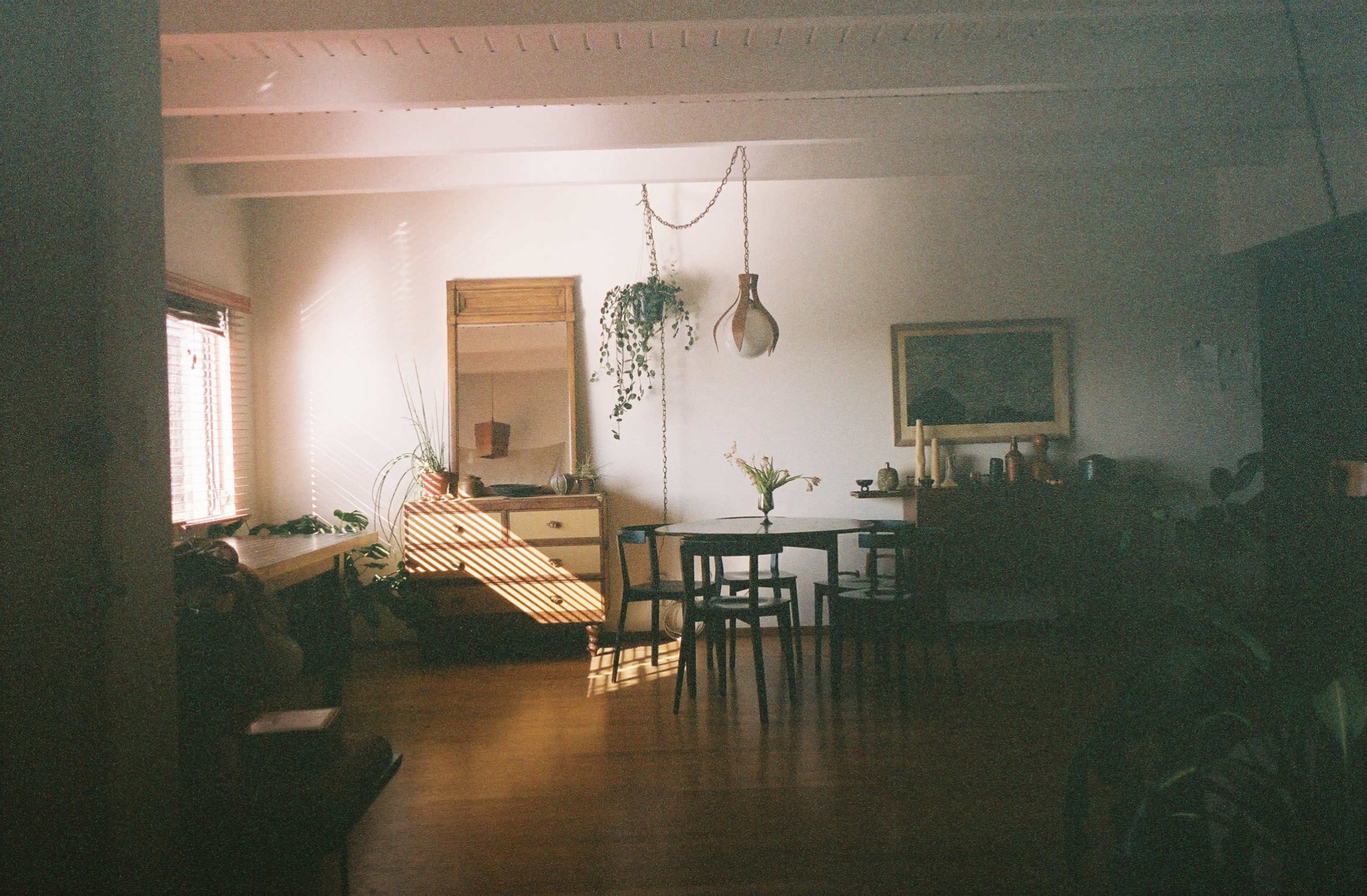 A dining area features a table surrounded by chairs, with sunlight filtering through a window and casting shadows on the wooden floor.