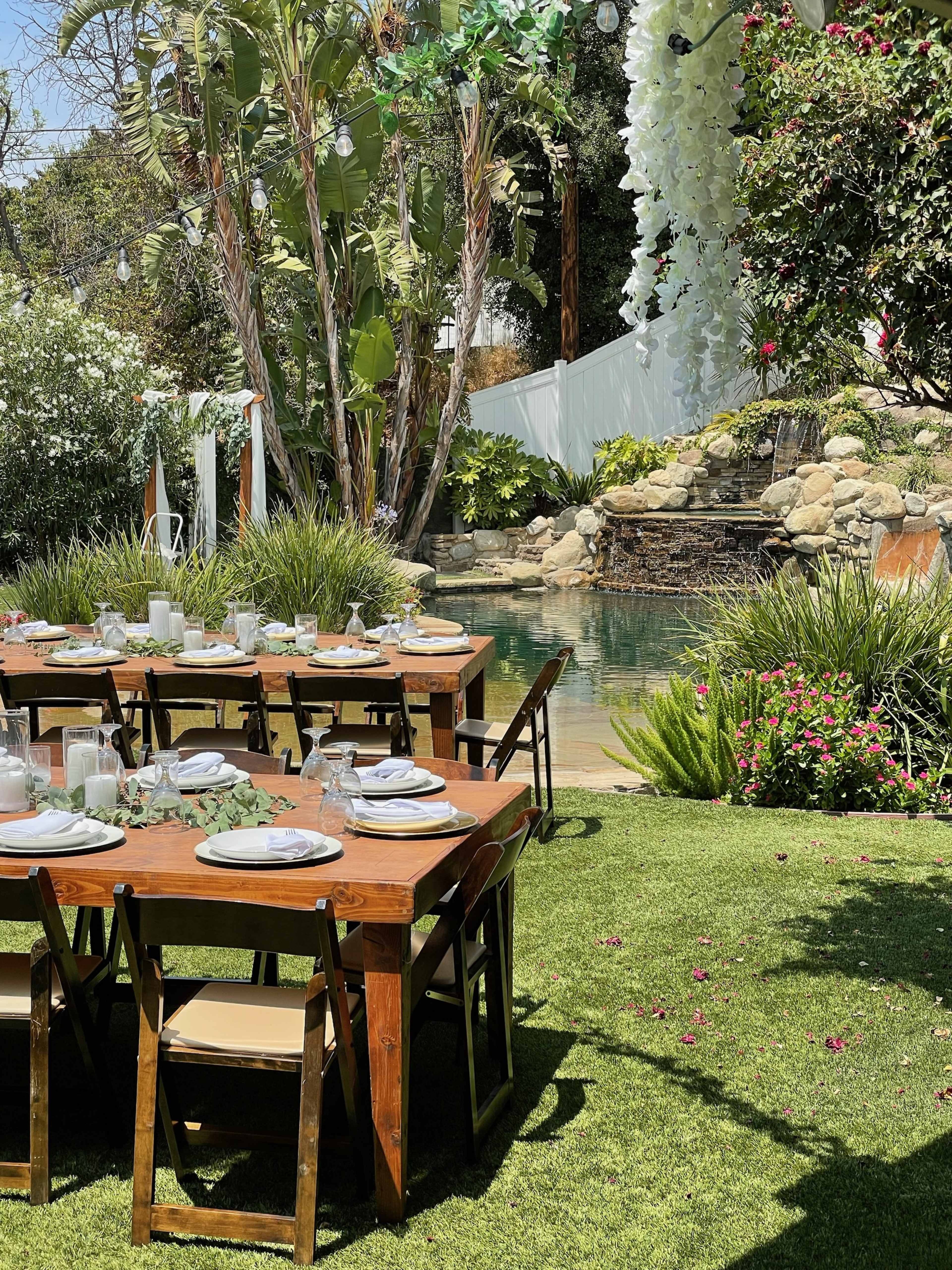 A wooden dining table is set up outside near a pool surrounded by greenery and flowers.