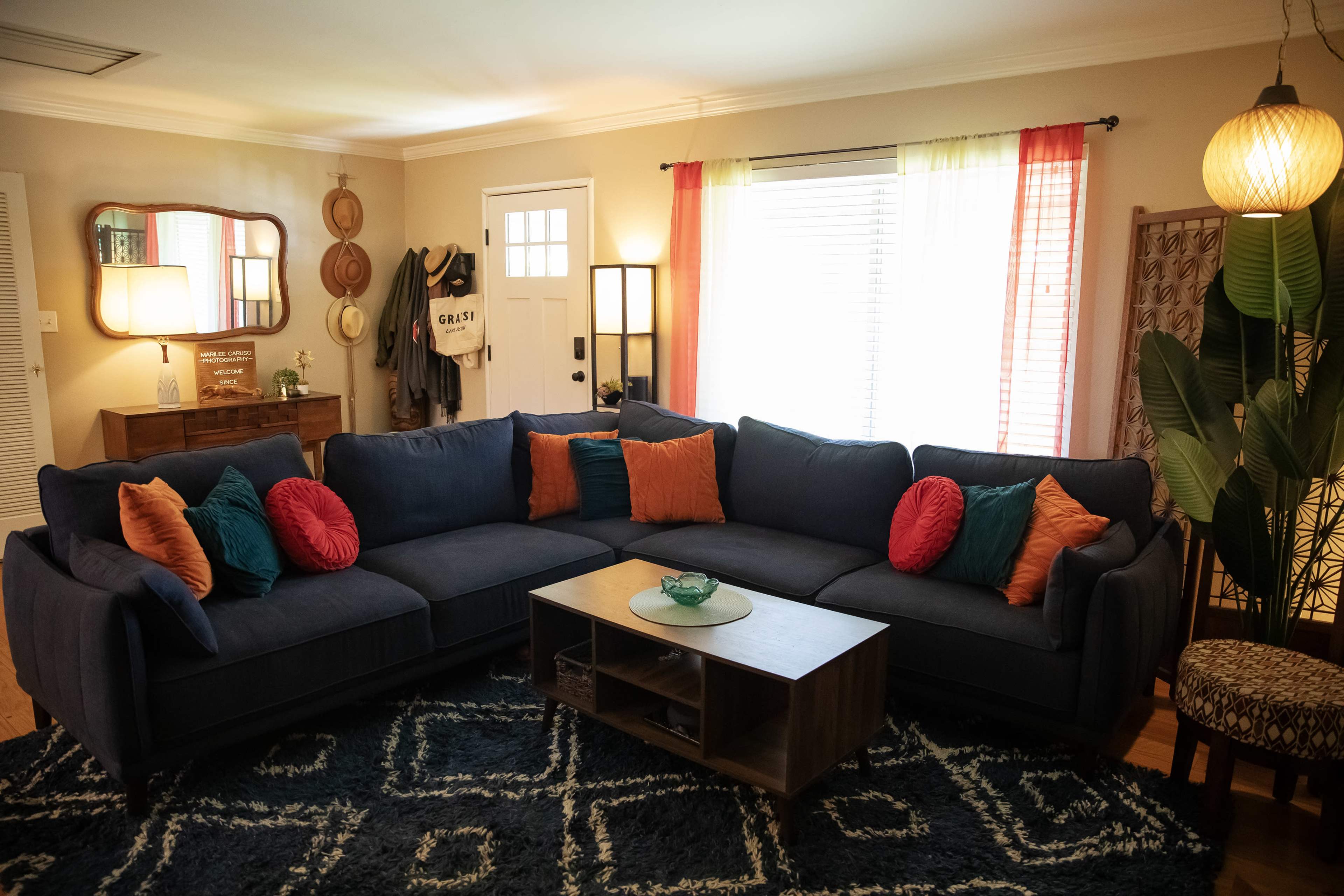 A spacious living room features a large blue sectional sofa with colorful cushions, a wooden coffee table, and a mix of decorative elements, including a floor lamp and plants.