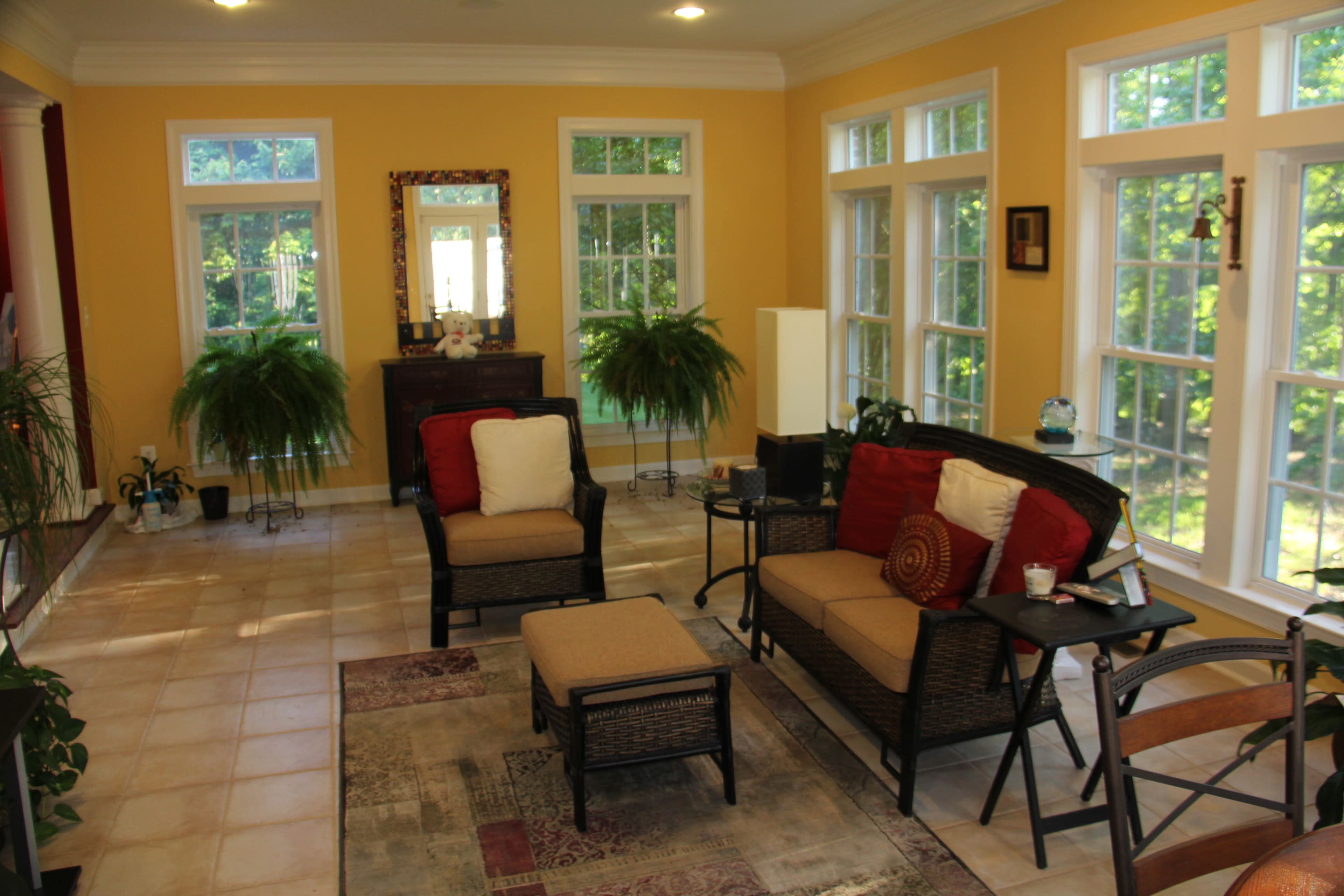 A sunny room with yellow walls, featuring wicker furniture, large windows, and several indoor plants.