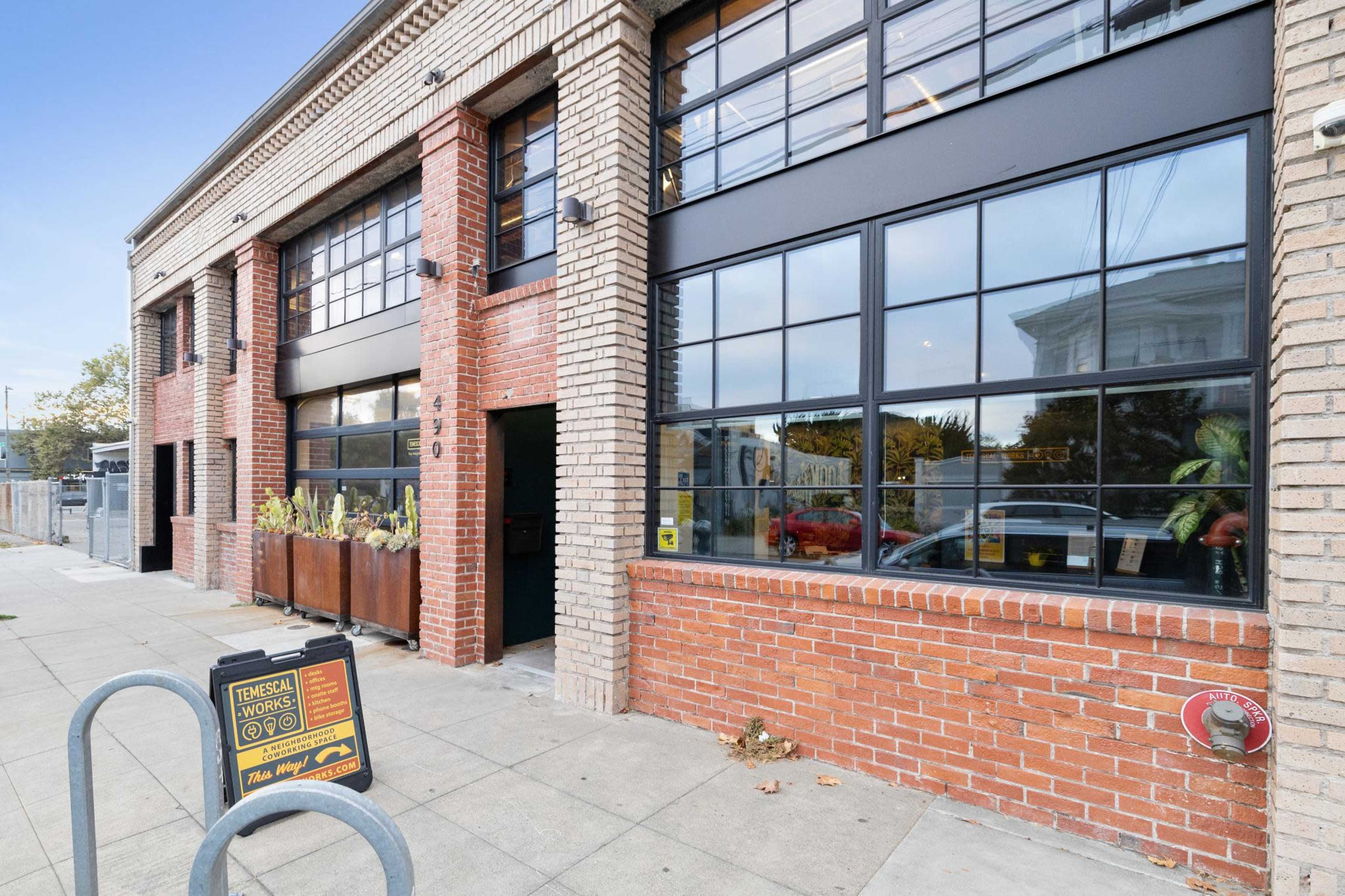 The image shows a brick building with large windows and a sign outside, located on a sidewalk.
