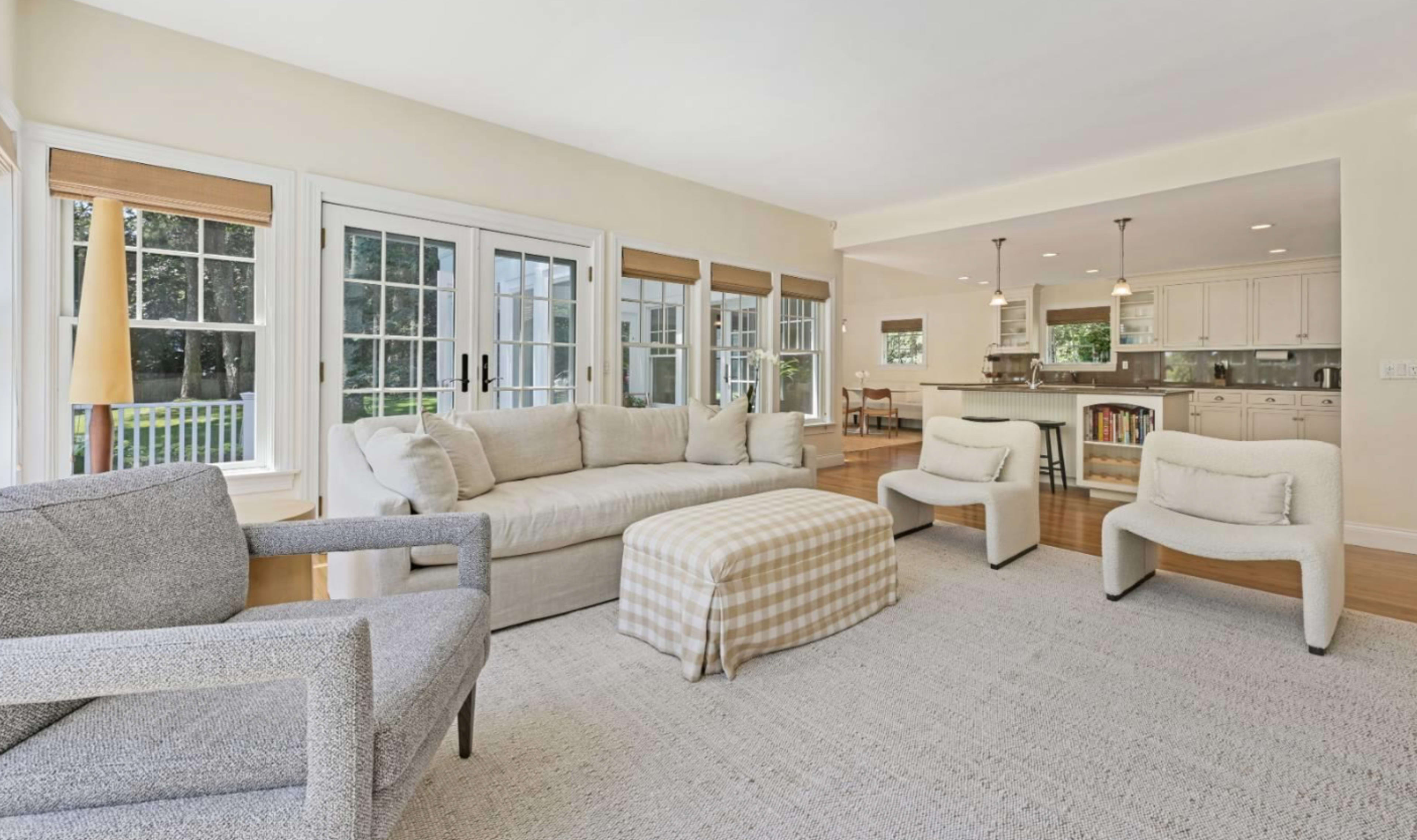 A well-lit living area featuring a beige sofa, two chairs, and a checkered ottoman, with large windows providing a view of the outdoors.