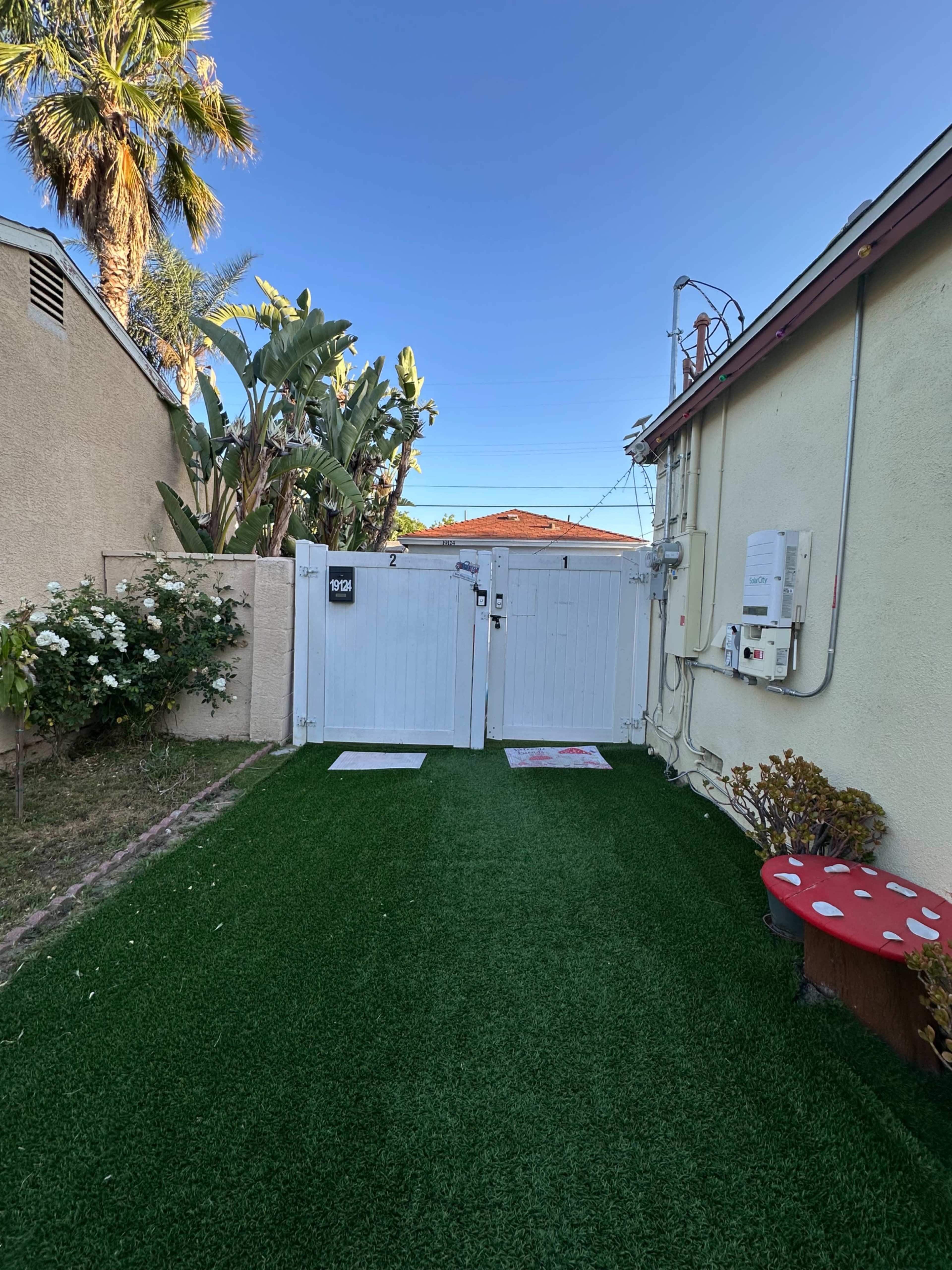 The image shows a narrow pathway lined with artificial grass leading to a white gate, flanked by plants and a small decorative mushroom statue.